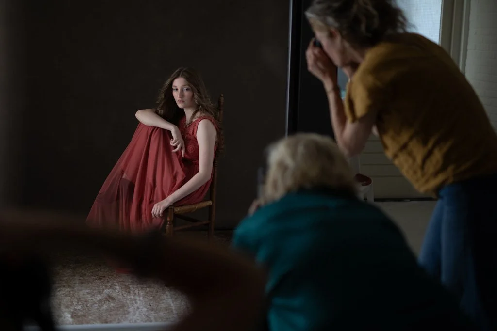 Een fotoshoot in een studio met een jonge vrouw in een rode jurk op de achtergrond en een fotograaf en assistent op de voorgrond.