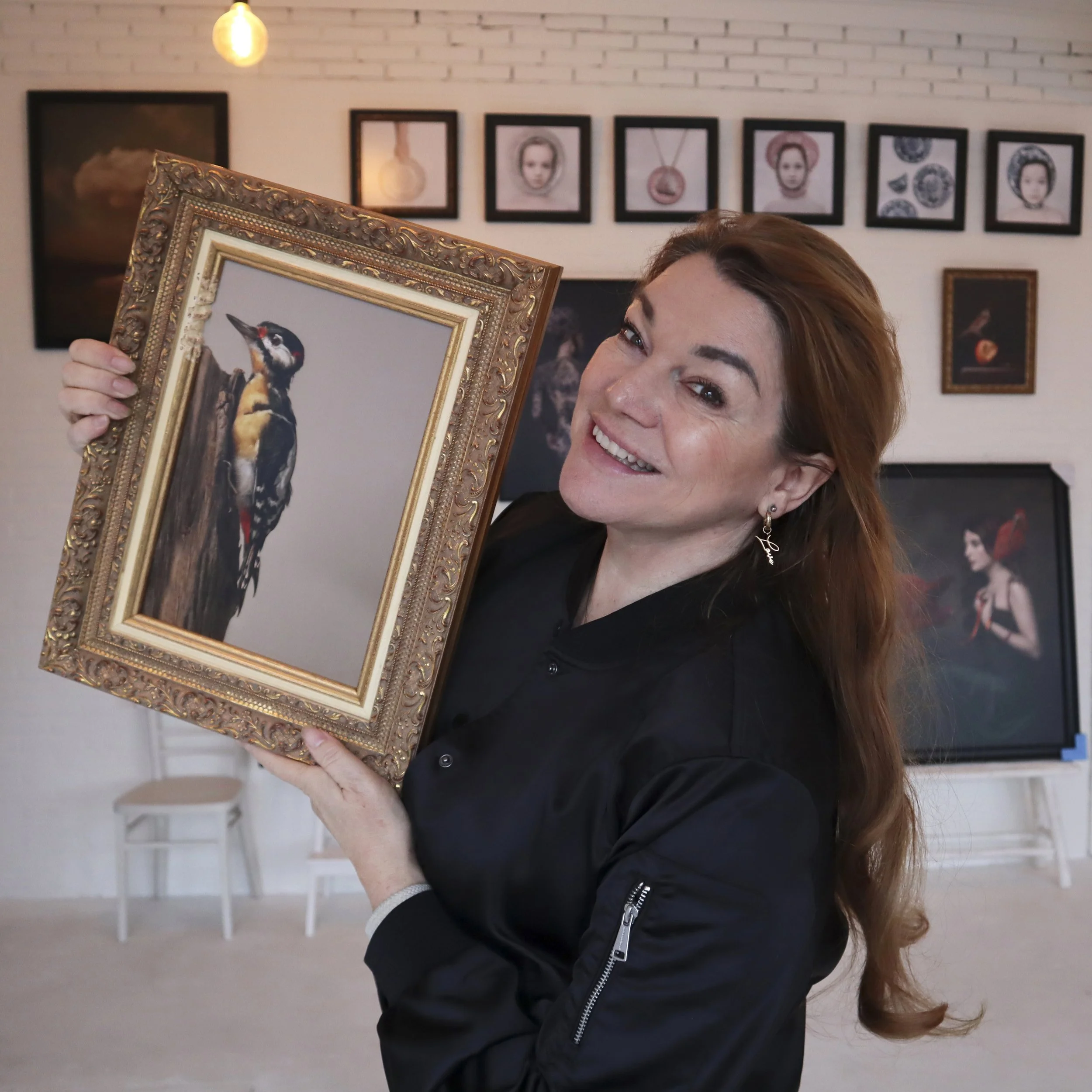 Vrouw met bruin haar glimlachend en een schilderij van een specht vasthoudend, binnen in een galerie met witte muren en foto's aan de muur op de achtergrond.