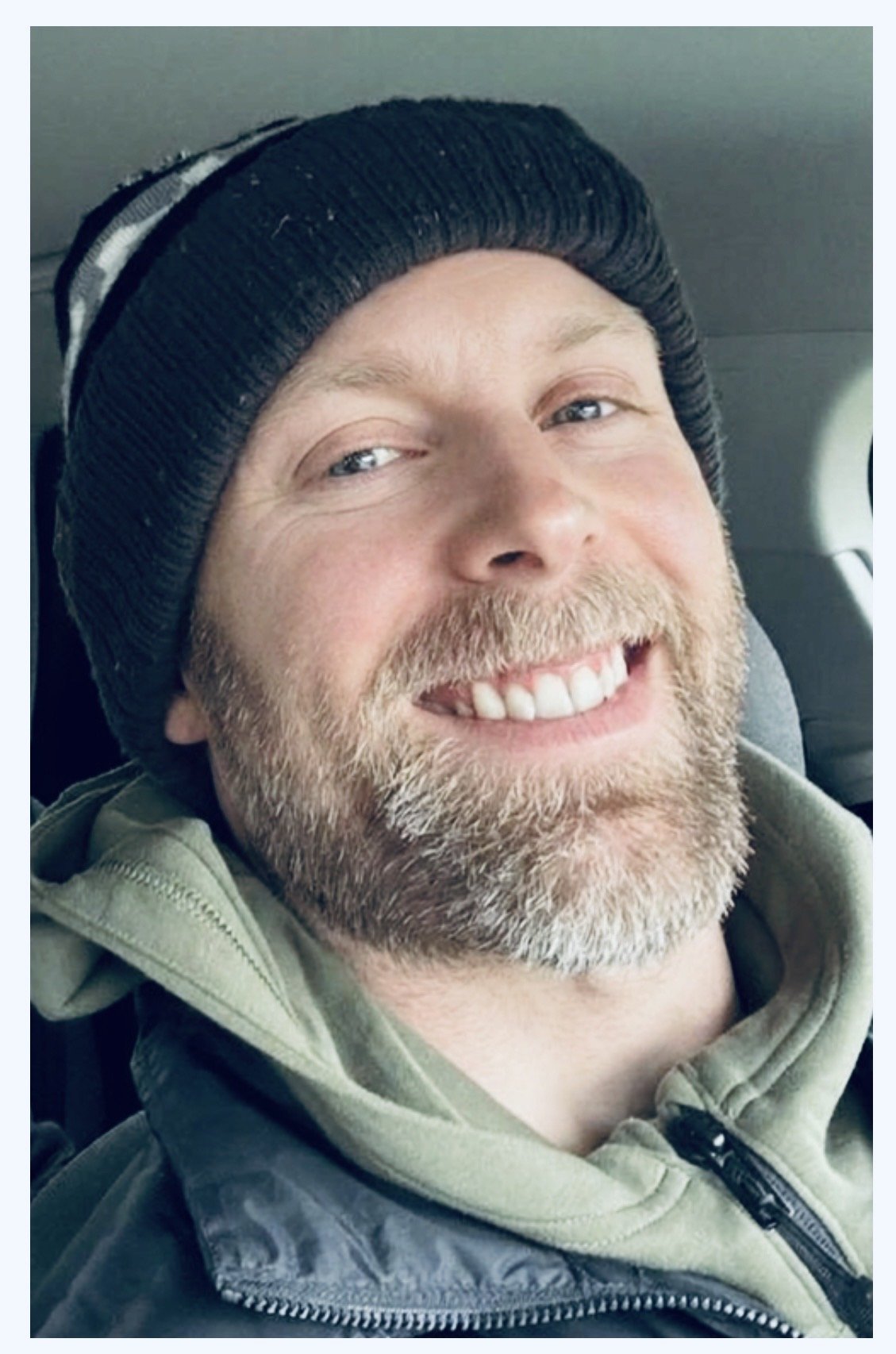 A smiling man with a beard wearing a black knit beanie, a green jacket, and a beige hoodie.