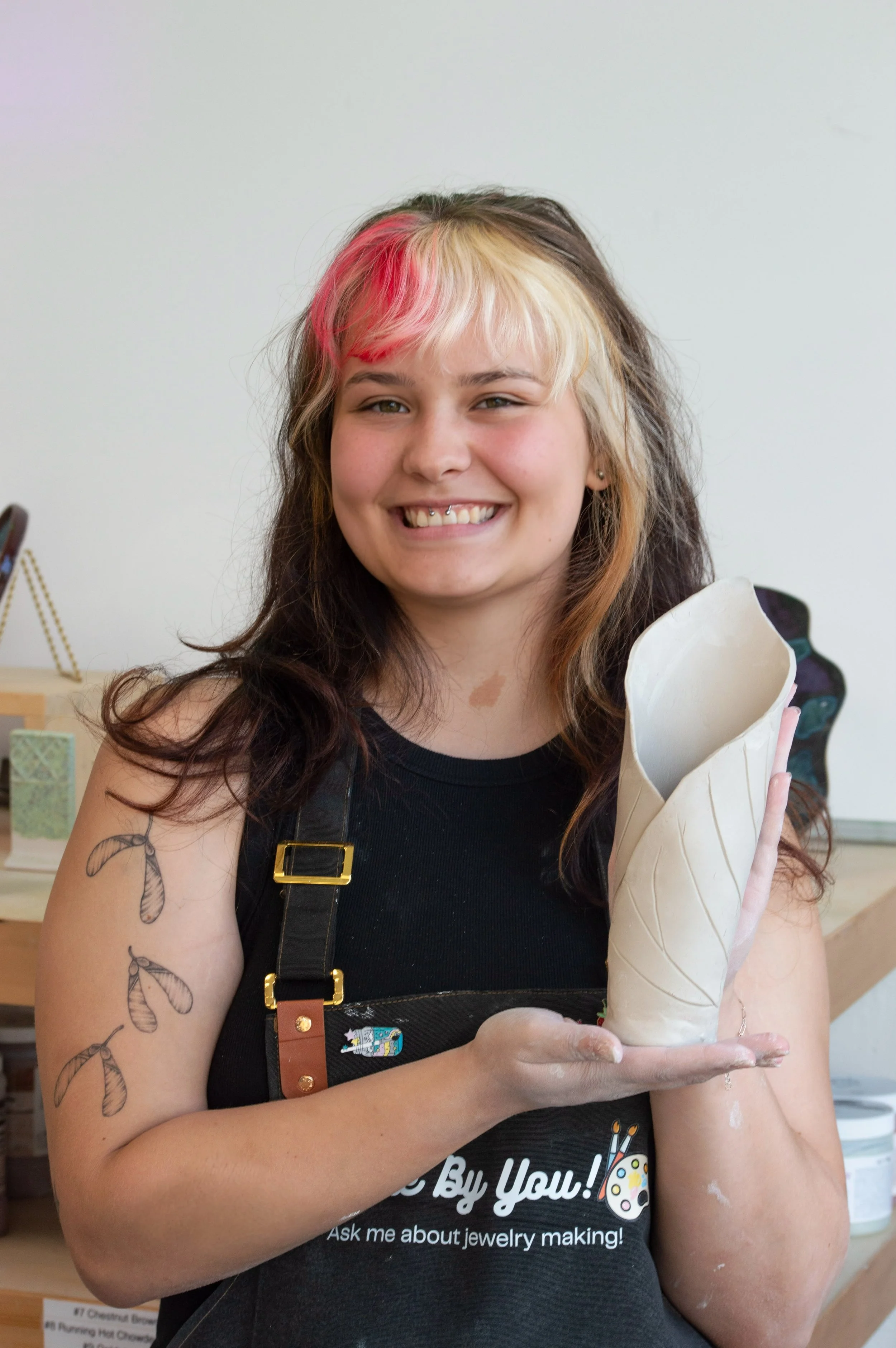 A young woman with blonde and brown hair, some pink highlights, and tattoos on her arm, smiling and holding a ceramic hand sculpture in a jewelry making class.