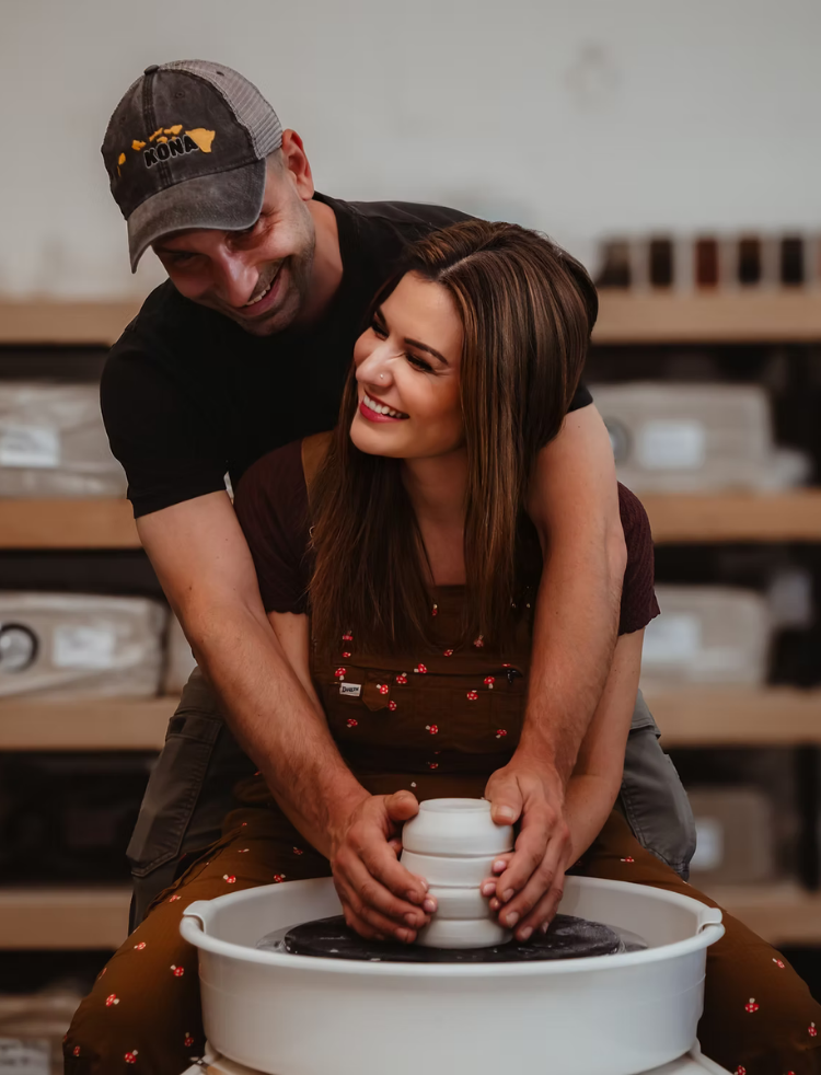 A man and woman enjoying a pottery-making activity together, with the man guiding the woman's hands on a pottery wheel, both smiling.