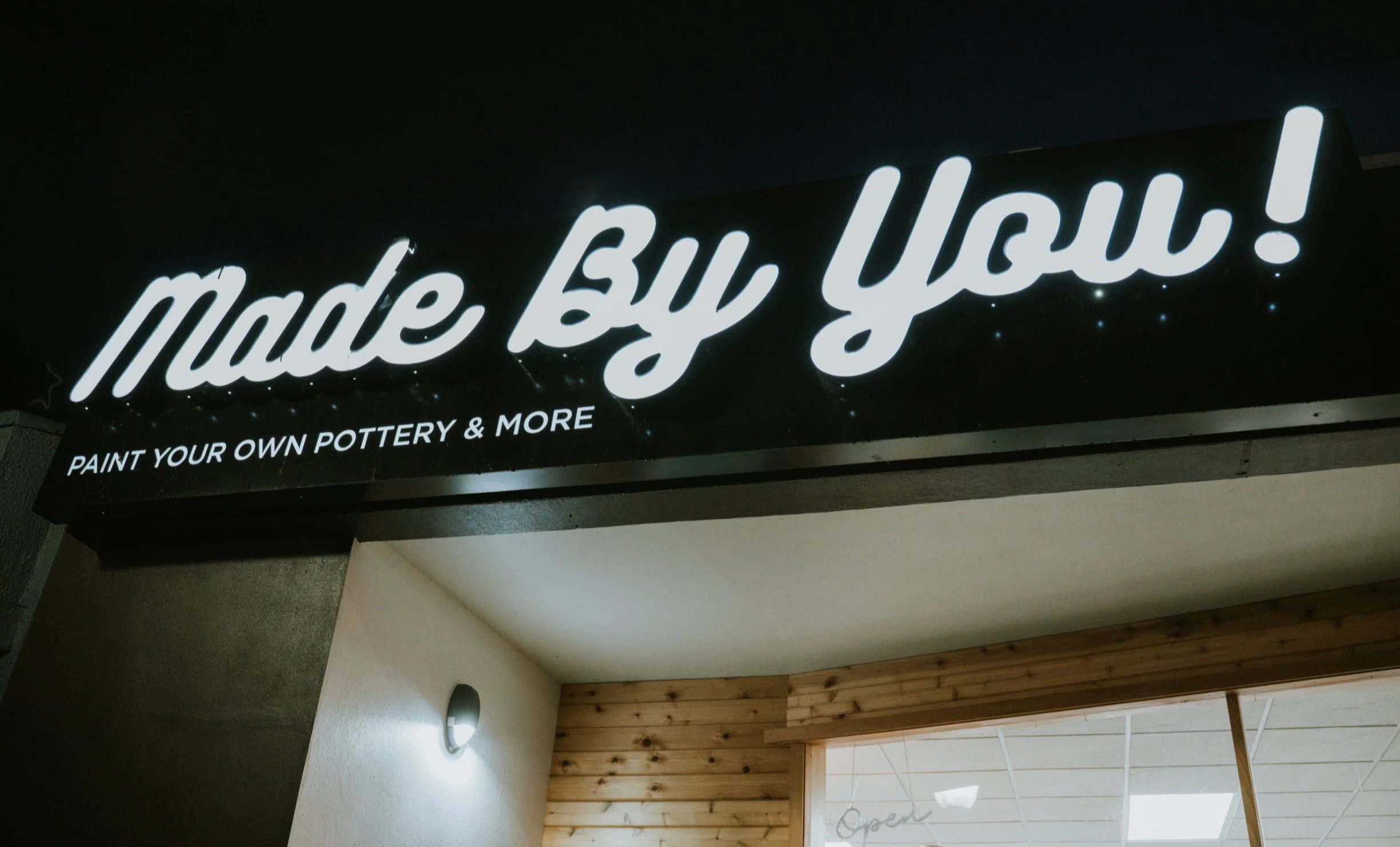 An illuminated sign that reads 'Made By You!' with smaller text below that says 'Paint Your Own Pottery & More'.