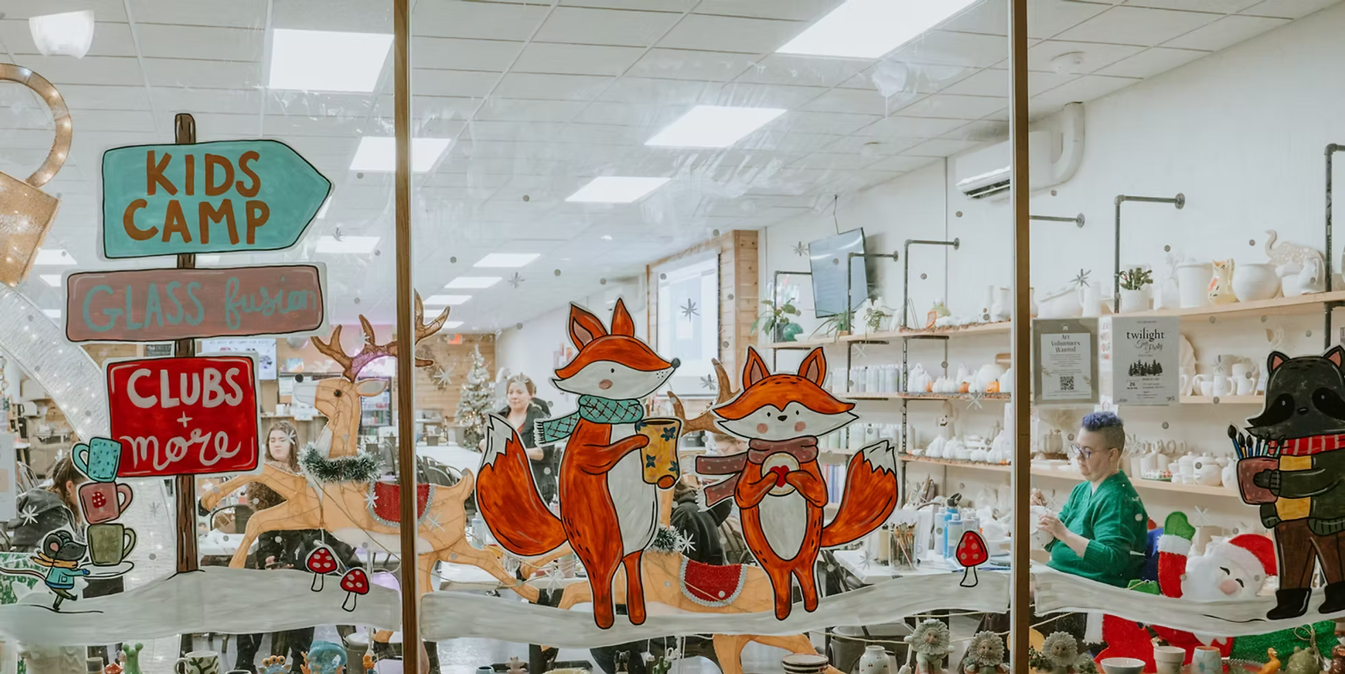 Decorative storefront window with cartoon fox and raccoon characters, Christmas trees, and holiday signs advertising kids camp, glass fusion, clubs, and more. Inside, people are working on crafts amidst shelves of holiday pottery and decorations.