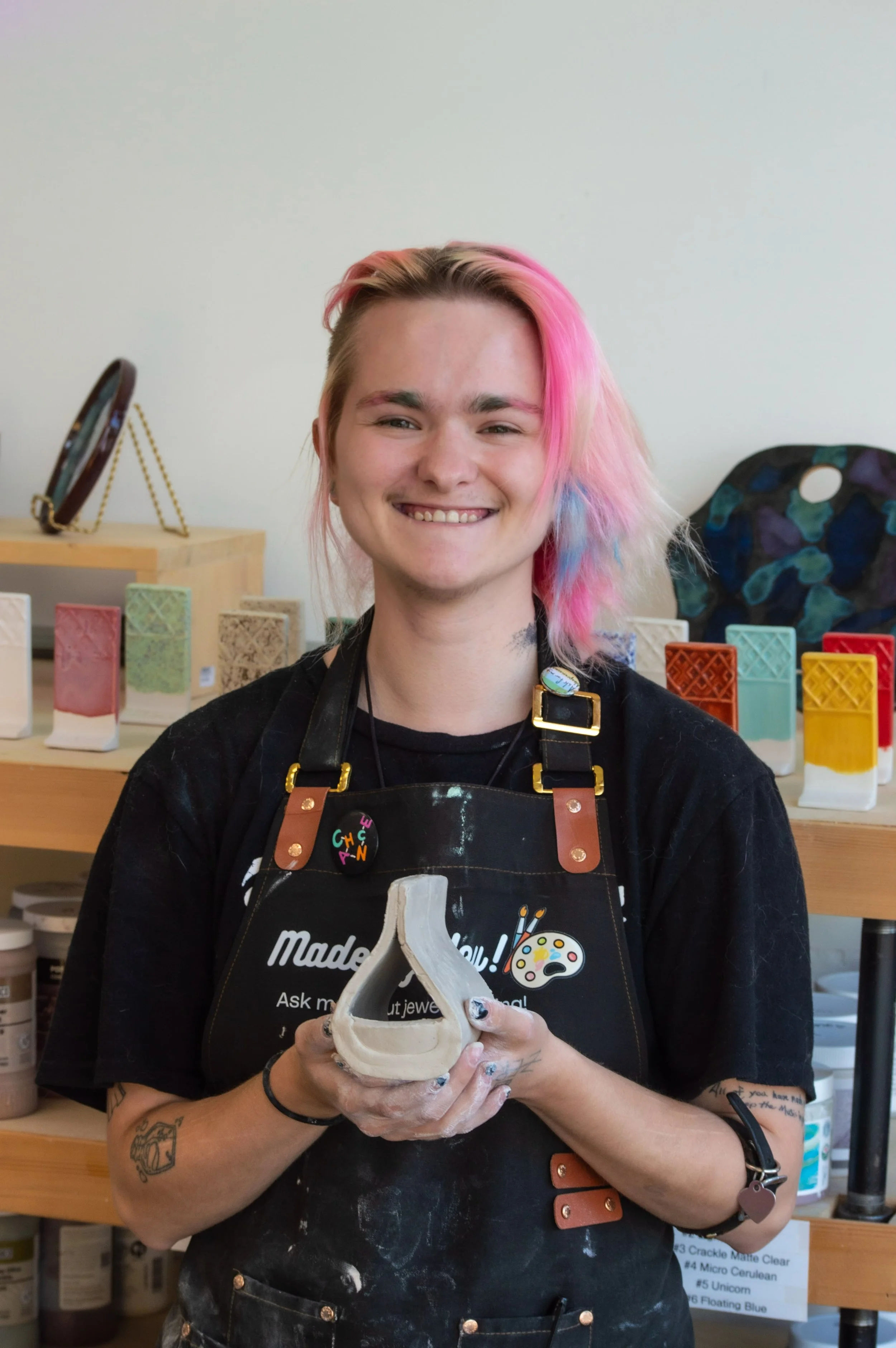 A smiling person with pink and blonde hair, wearing a black apron and holding a ceramic skull, standing in a workspace with colorful ceramic pieces in the background.