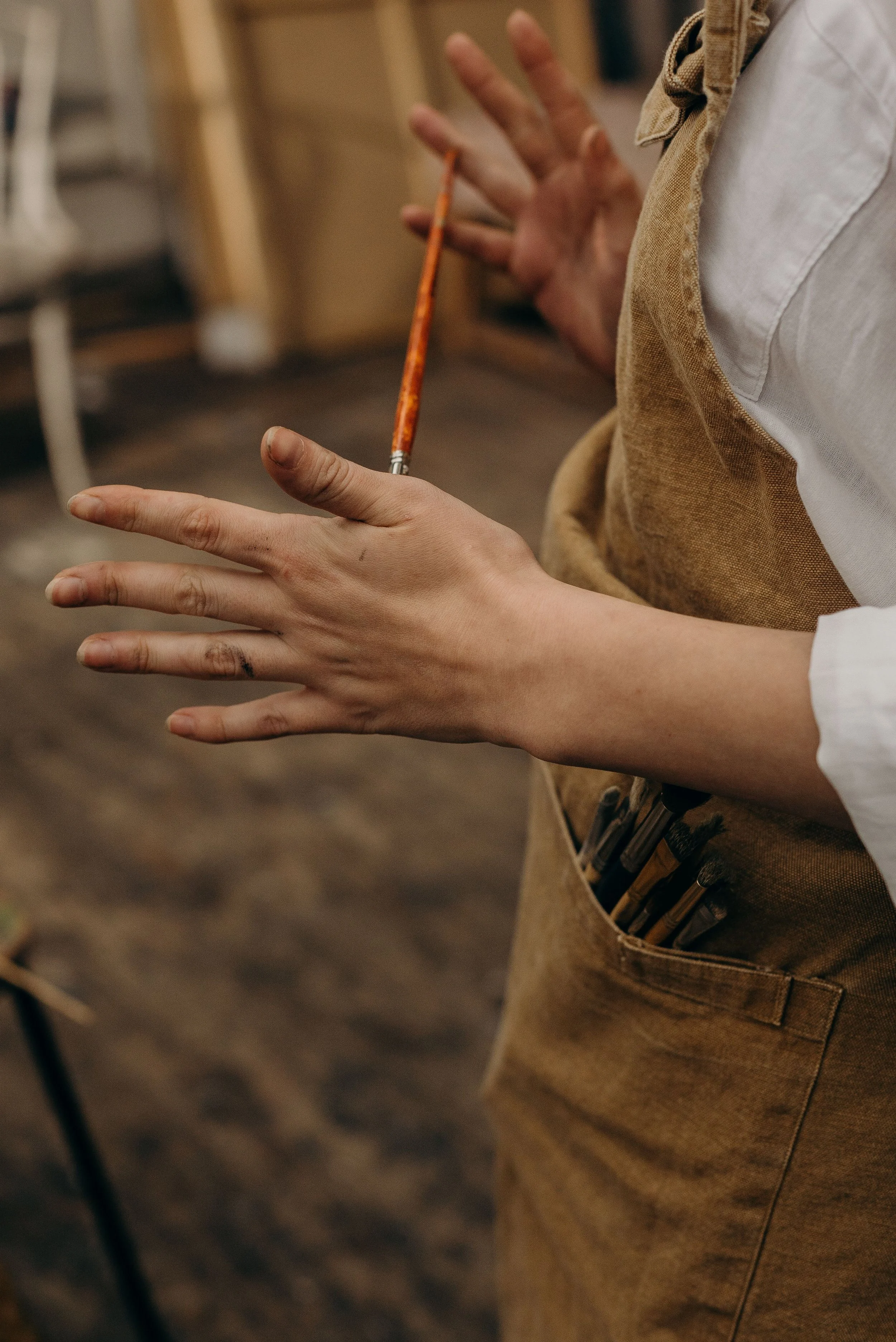 Close-up of a person's hands and torso. The person is wearing a beige apron with visible pockets, holding a paintbrush in one hand, in what appears to be a workshop or art studio.
