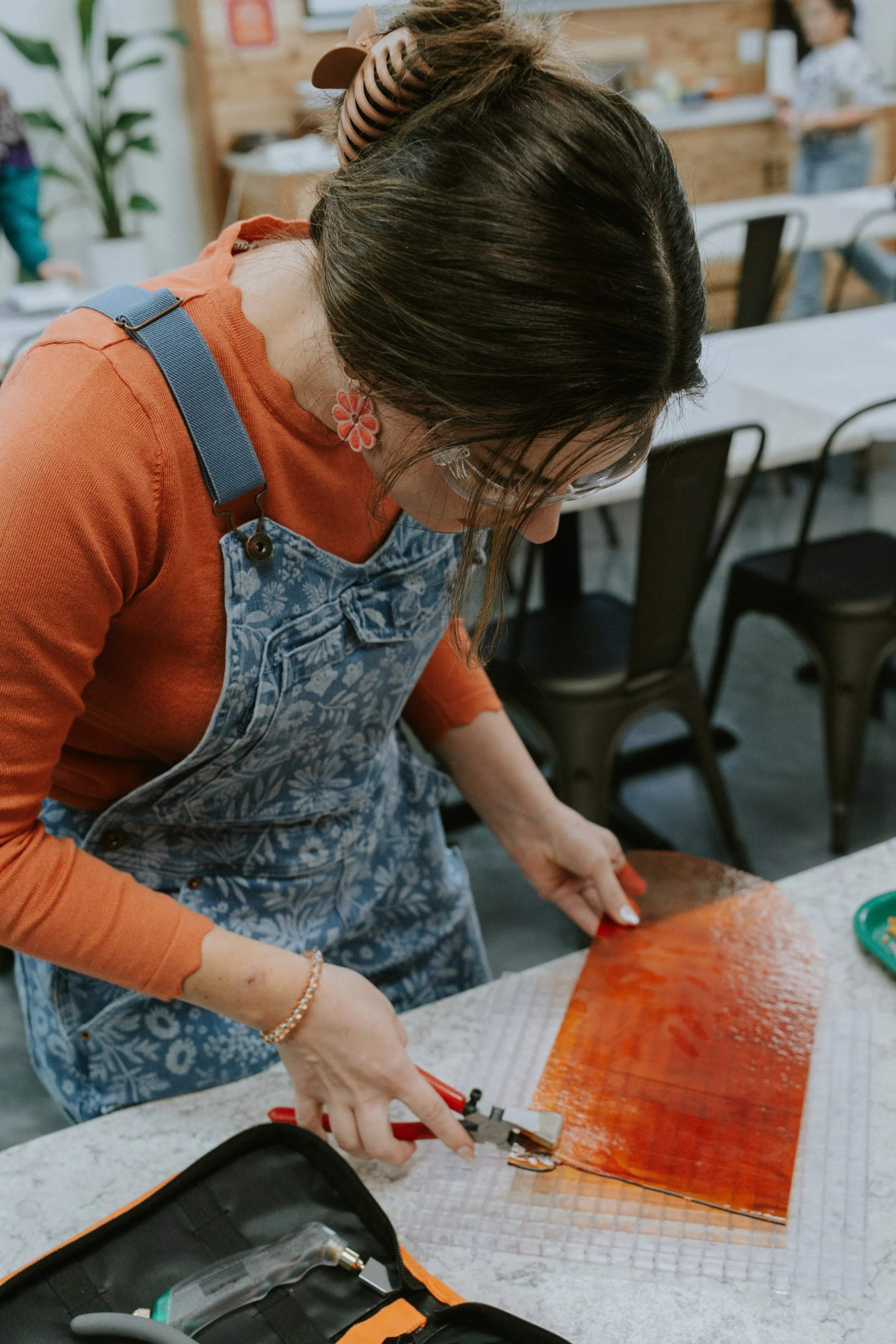A woman with dark hair, glasses, and floral earrings is cutting a large piece of salmon with scissors on a cutting board in a kitchen or workshop setting. Other people are visible in the background.