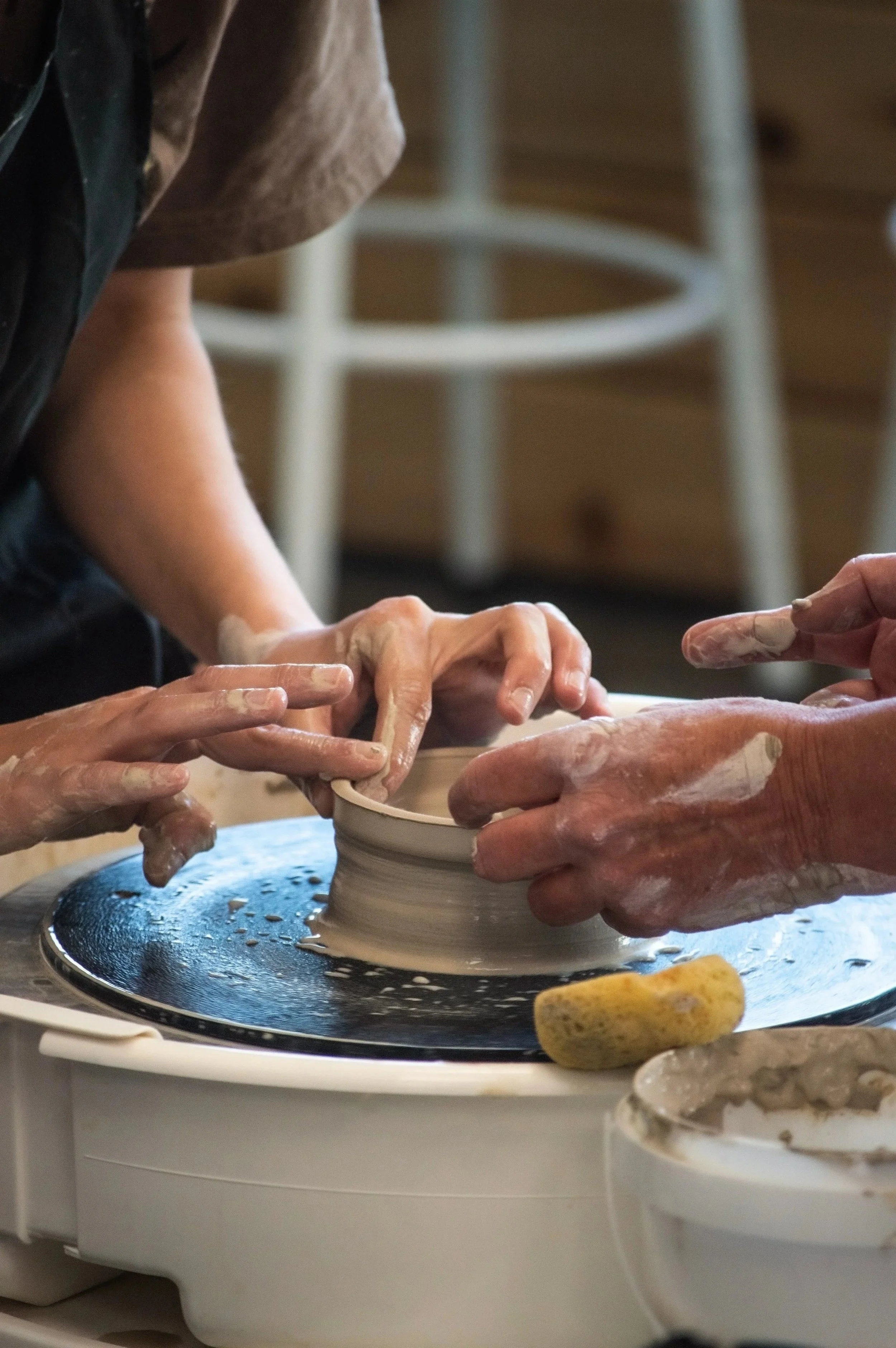 Close-up of hands shaping a clay pottery on a pottery wheel, with a blurred background of a chair and other pottery items.
