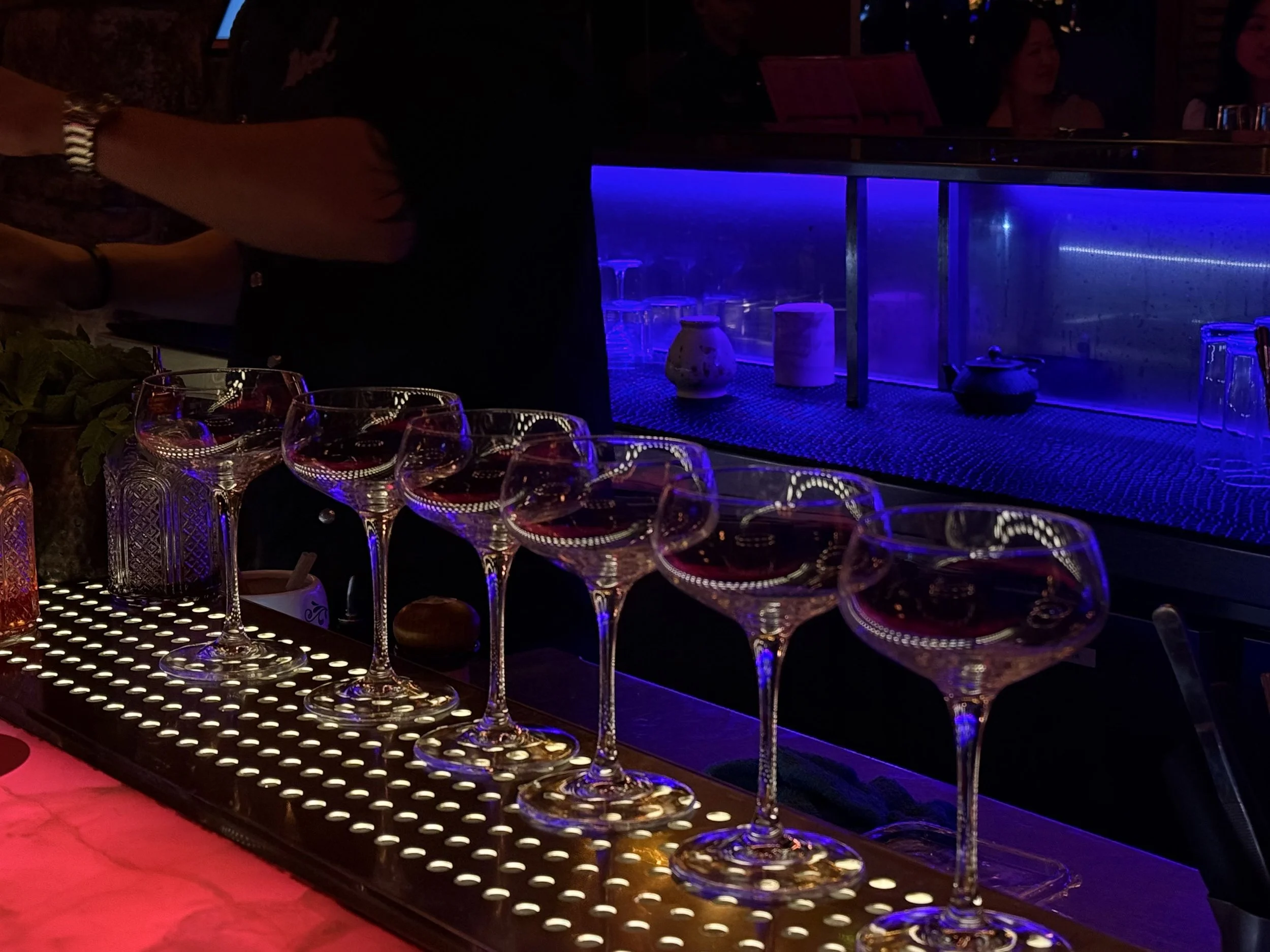 A bar counter with several empty wine glasses lined up. In the background, there is a glass display with pottery and a glowing blue light. People are seated at the bar and in the background, though not clearly visible.
