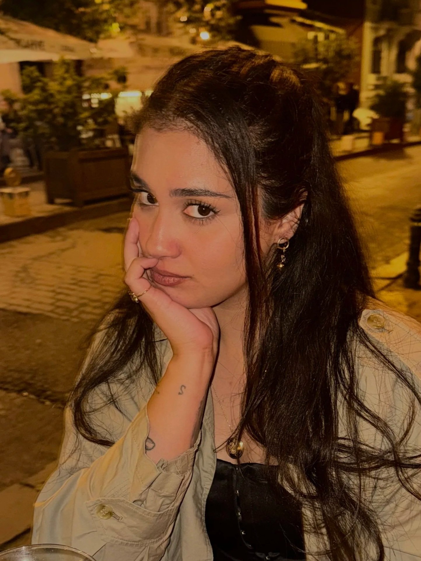 A young woman with long dark hair, wearing earrings and a ring, resting her face on her hand, sitting outdoors at night in an area with trees, benches, and a building behind her.