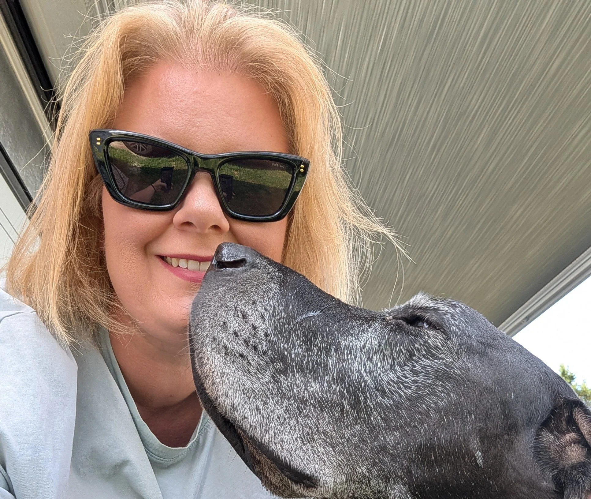 Lächelnde blonde Frau mit Sonnenbrille neben einem großen schwarz-grauen Hund im Freien