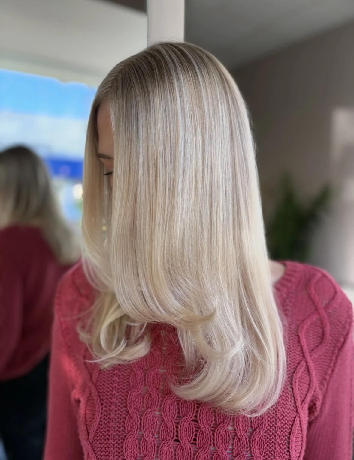Woman with shoulder-length blonde hair with soft waves, wearing a pink knit sweater, standing in front of a mirror in a well-lit room.