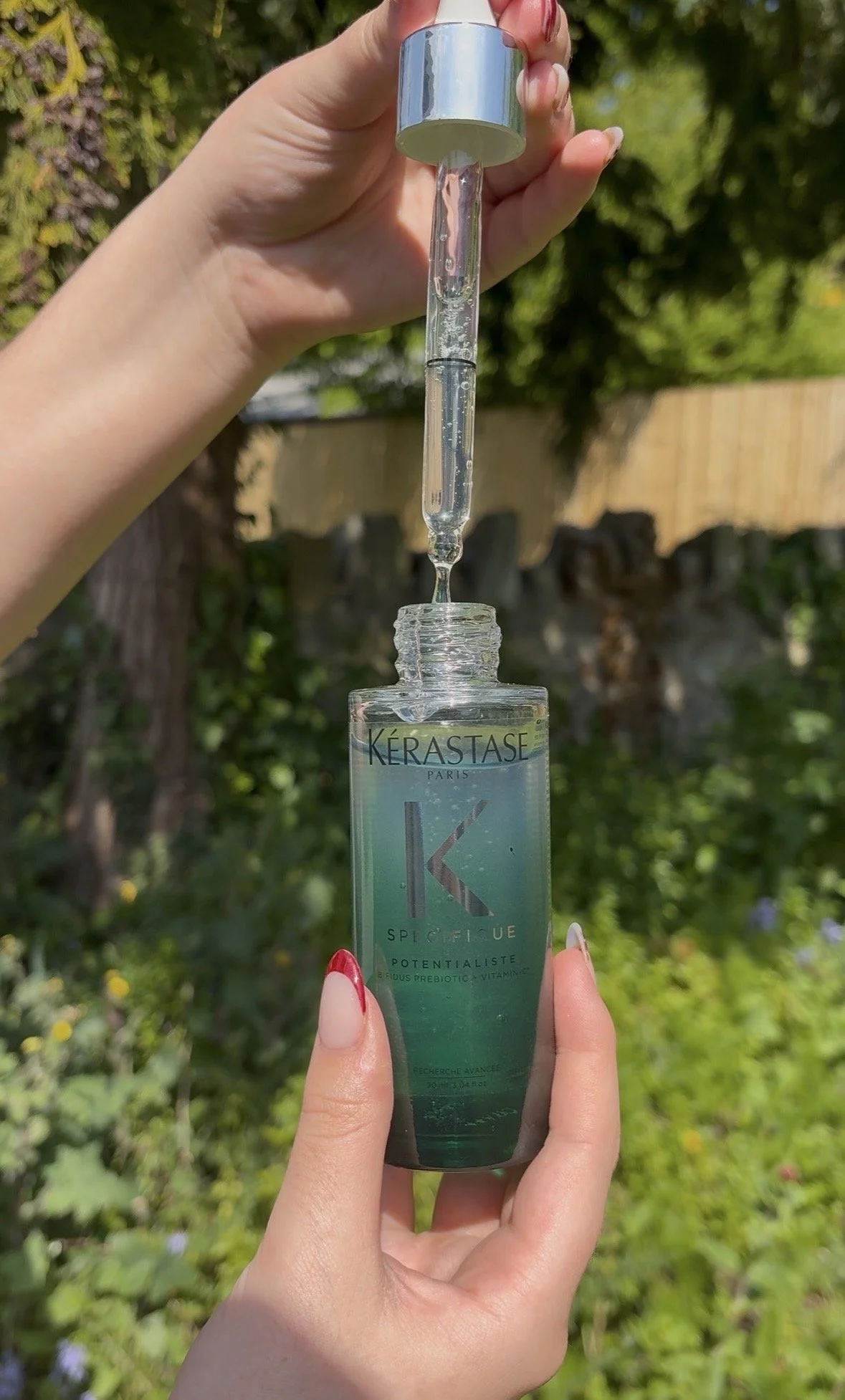Person holding a skincare serum bottle with a dropper, outdoors in a garden, green foliage in the background.