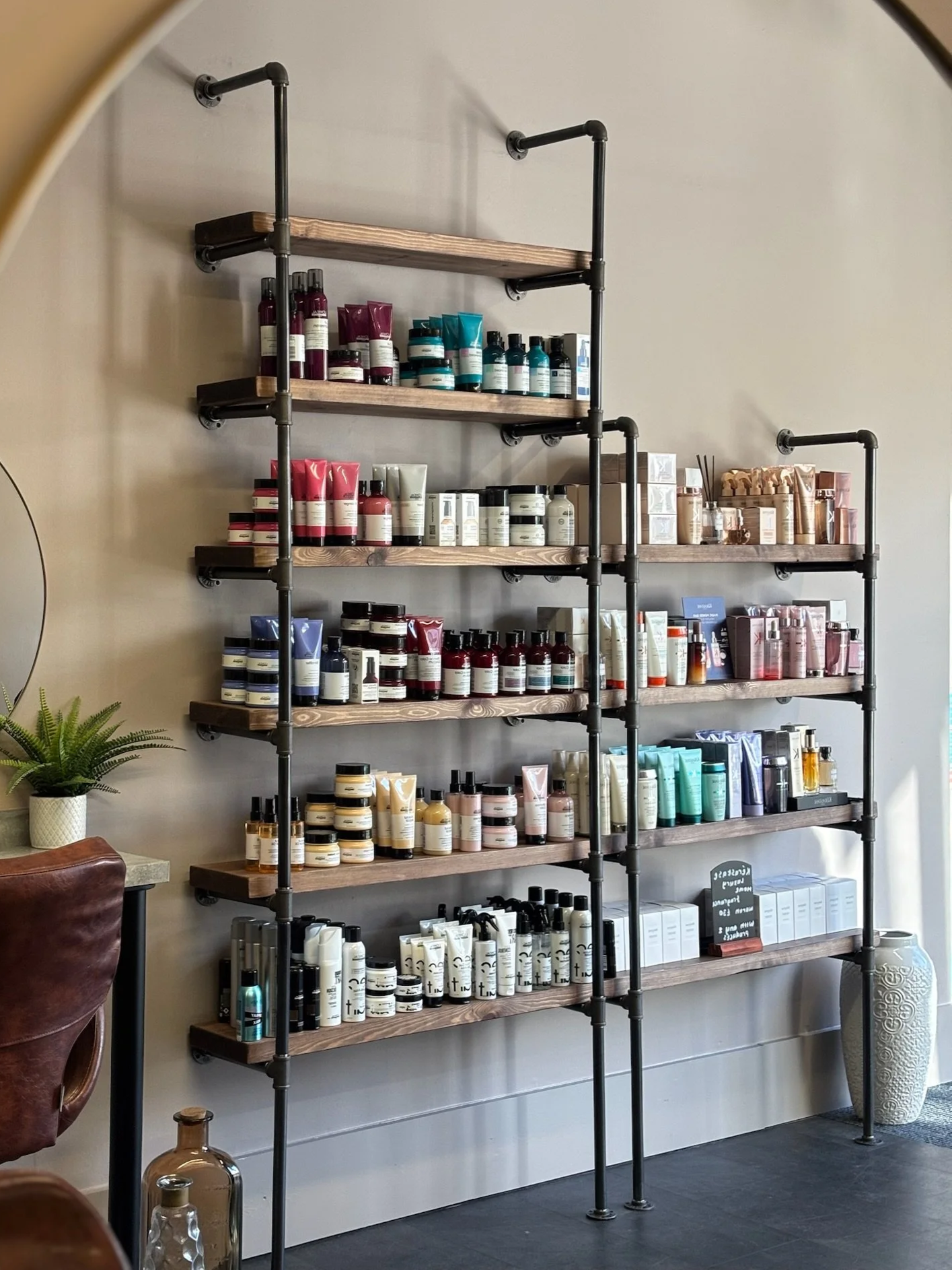 A black metal and wood shelving unit displaying various skincare and beauty products in bottles and tubes, arranged on six wooden shelves against a beige wall, with a green potted plant nearby.