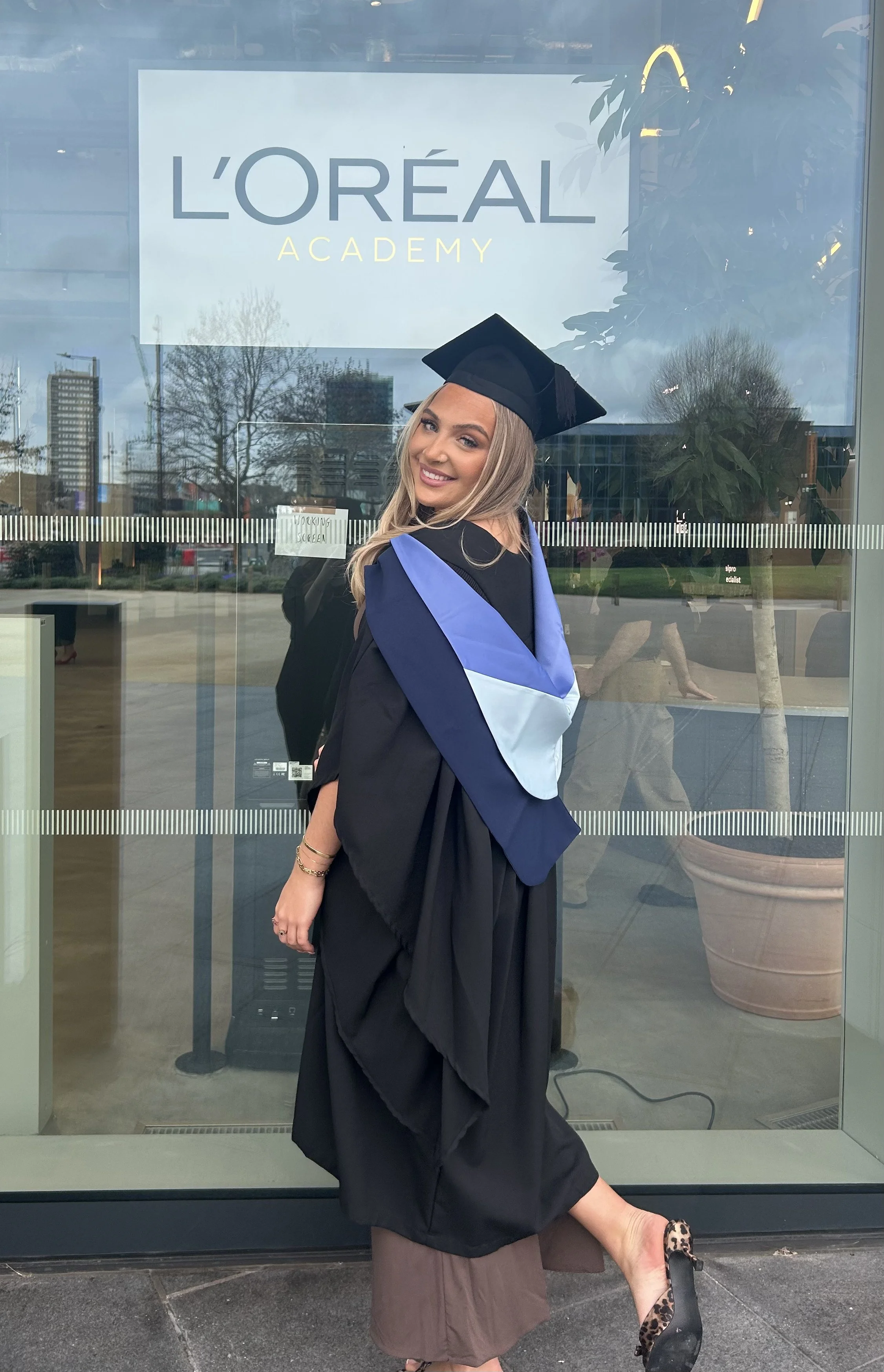 Graduate woman in cap and gown smiling, standing outside L'Oreal Academy with a window reflection of a cityscape and trees.