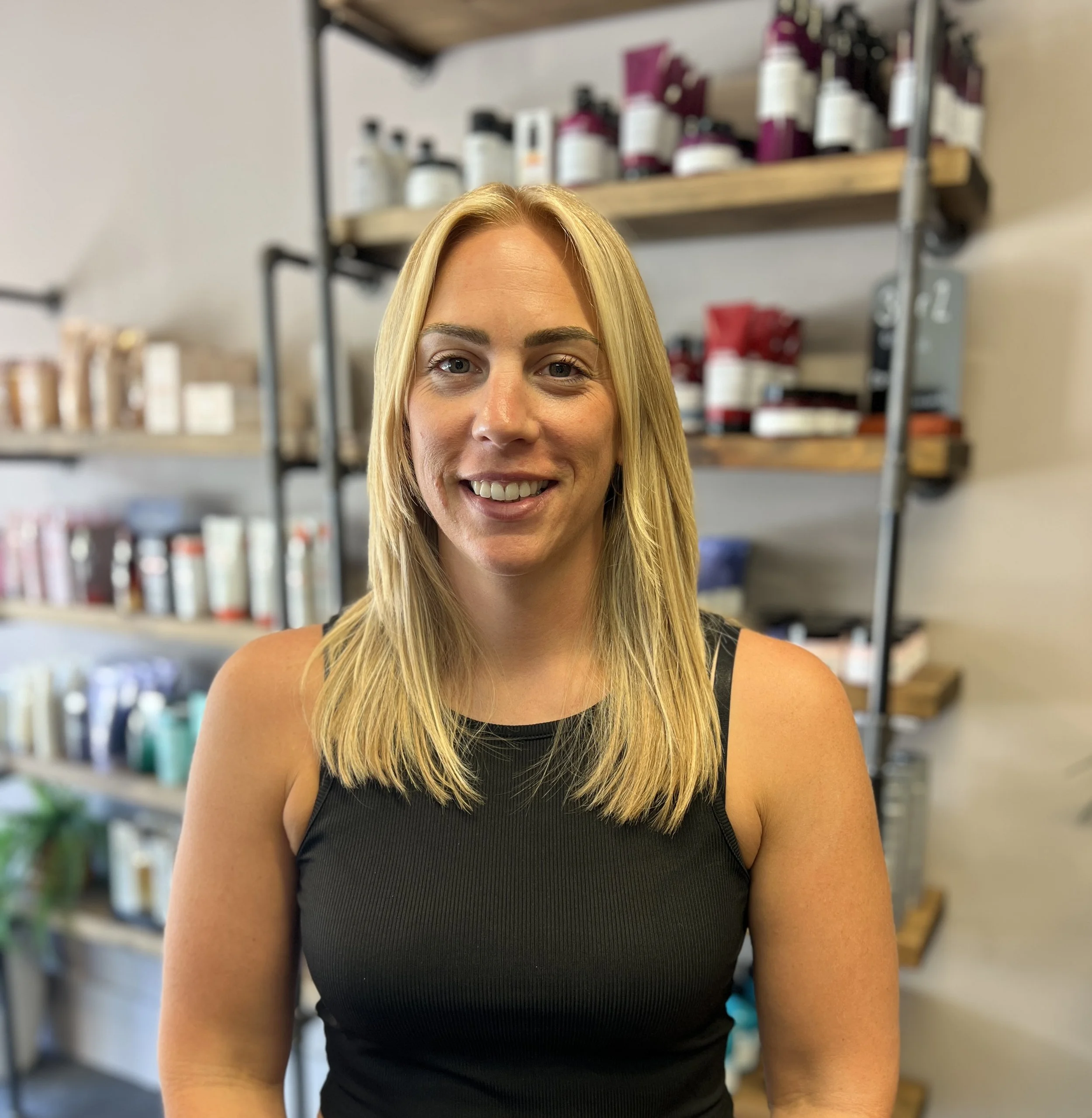 A smiling woman with blonde hair wearing a black sleeveless top, standing in front of shelves with hair and beauty products in a salon or store.