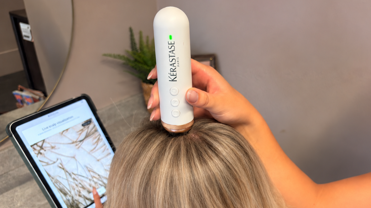 Person using a Kerastase scalp treatment device while reading a hair care website on a tablet.