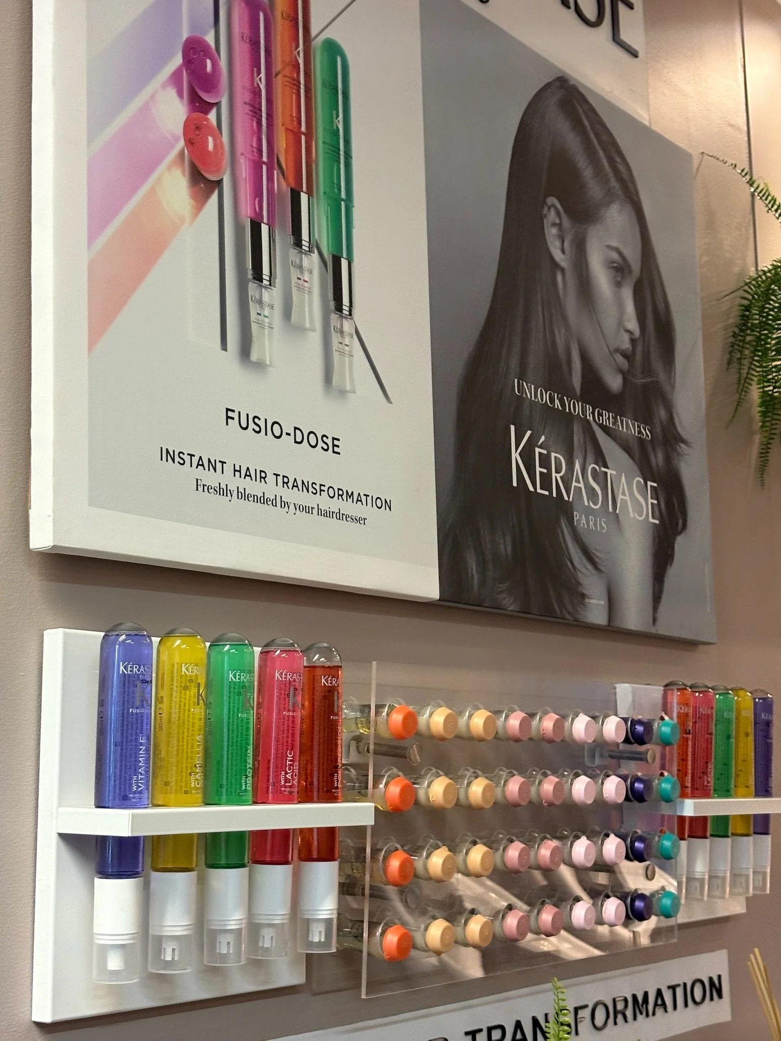 Display of Kérastase hair care products, including Fusio-Dose hair treatment tubes in various colors, with a large black and white poster of a woman with long, sleek hair and the text 'Unlock Your Greatness Kérastase Paris', and hair color samples on a shelf below.