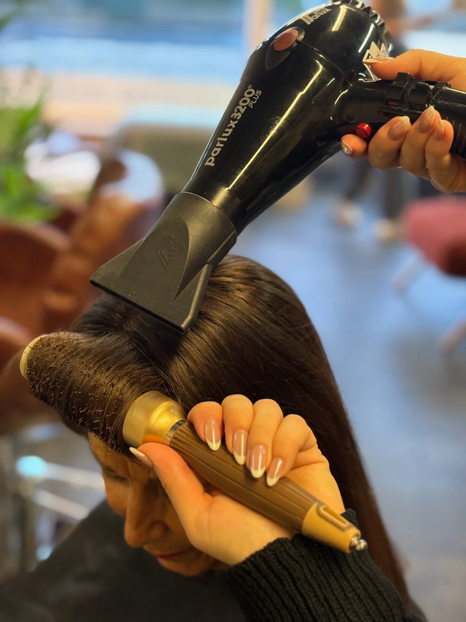 A person styling hair using a black and gold hairdryer in a salon.