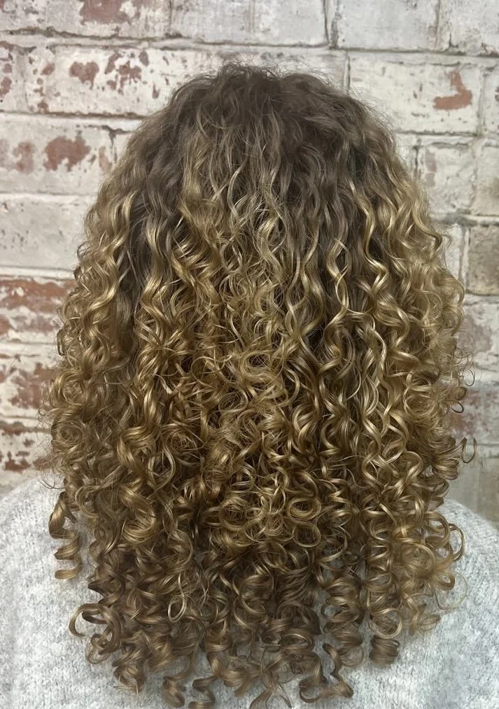 Back view of a person with shoulder-length, blonde, tightly curled hair against a brick wall.