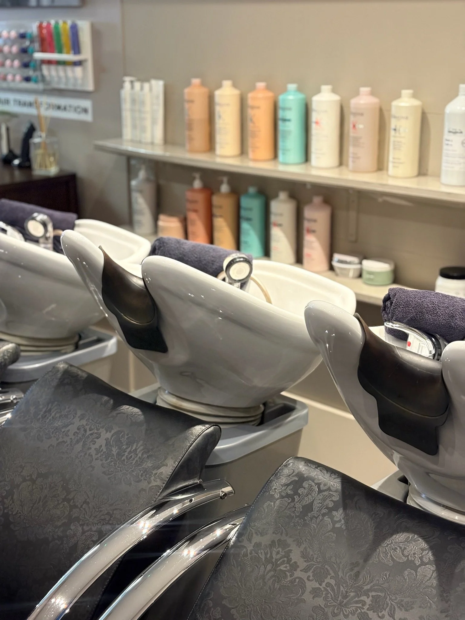 Salon shampoo stations with white ceramic sinks, dark patterned chairs, and shelves in the background stocked with various hair products.