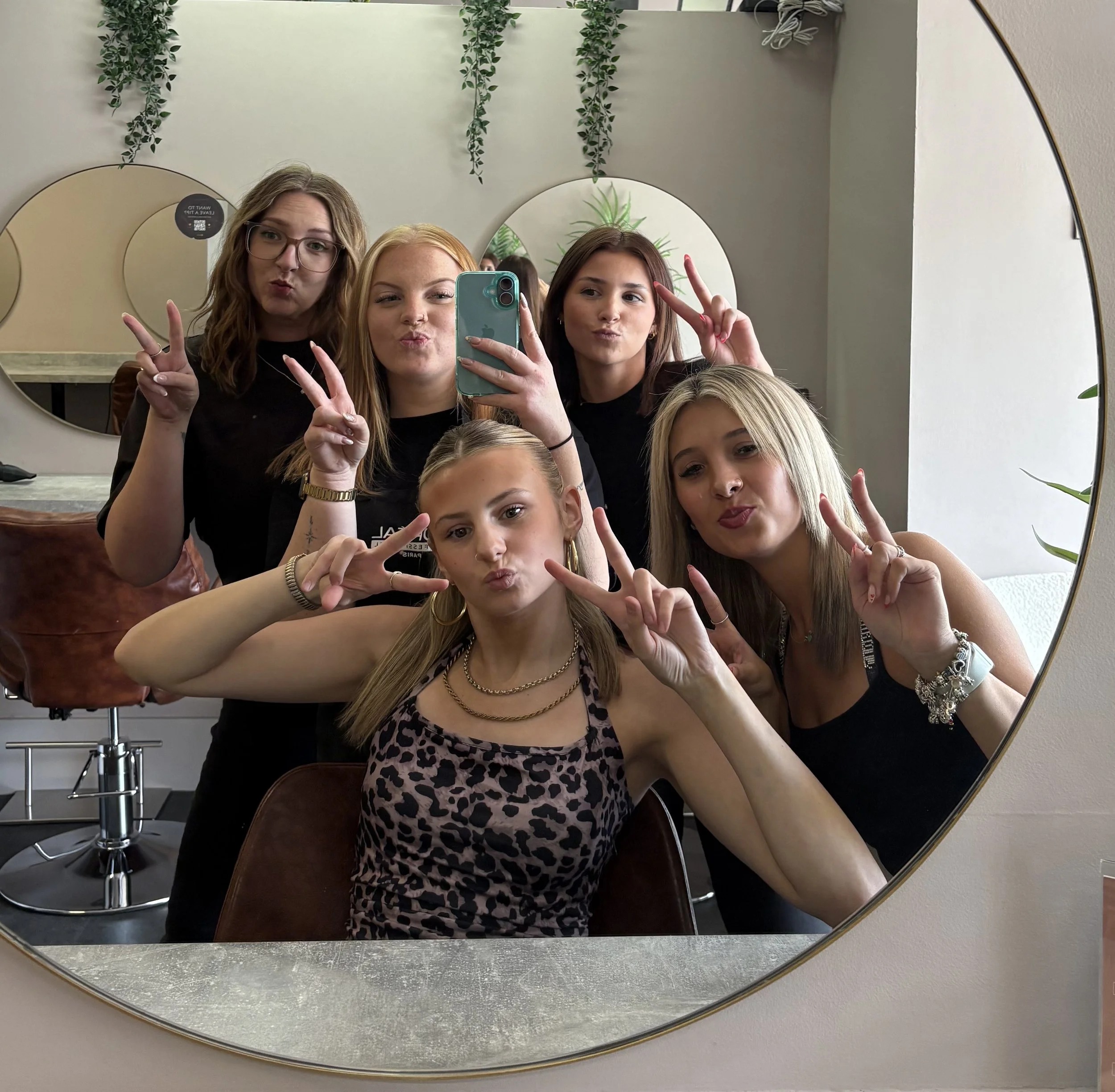 Group of women taking a selfie in a mirror, making peace signs and duck faces. The mirror reflects part of a room with hanging greenery and plants.