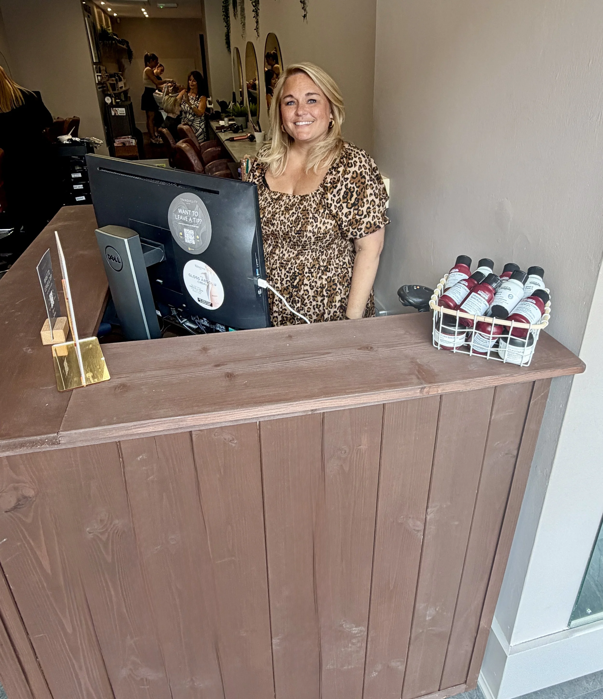 A woman with blonde hair smiling at a reception desk in a salon or beauty shop. Behind her, there are mirrors and clients getting services, with hairdressers attending to them. The reception desk has a computer, printed signs, and bottles of hair products.