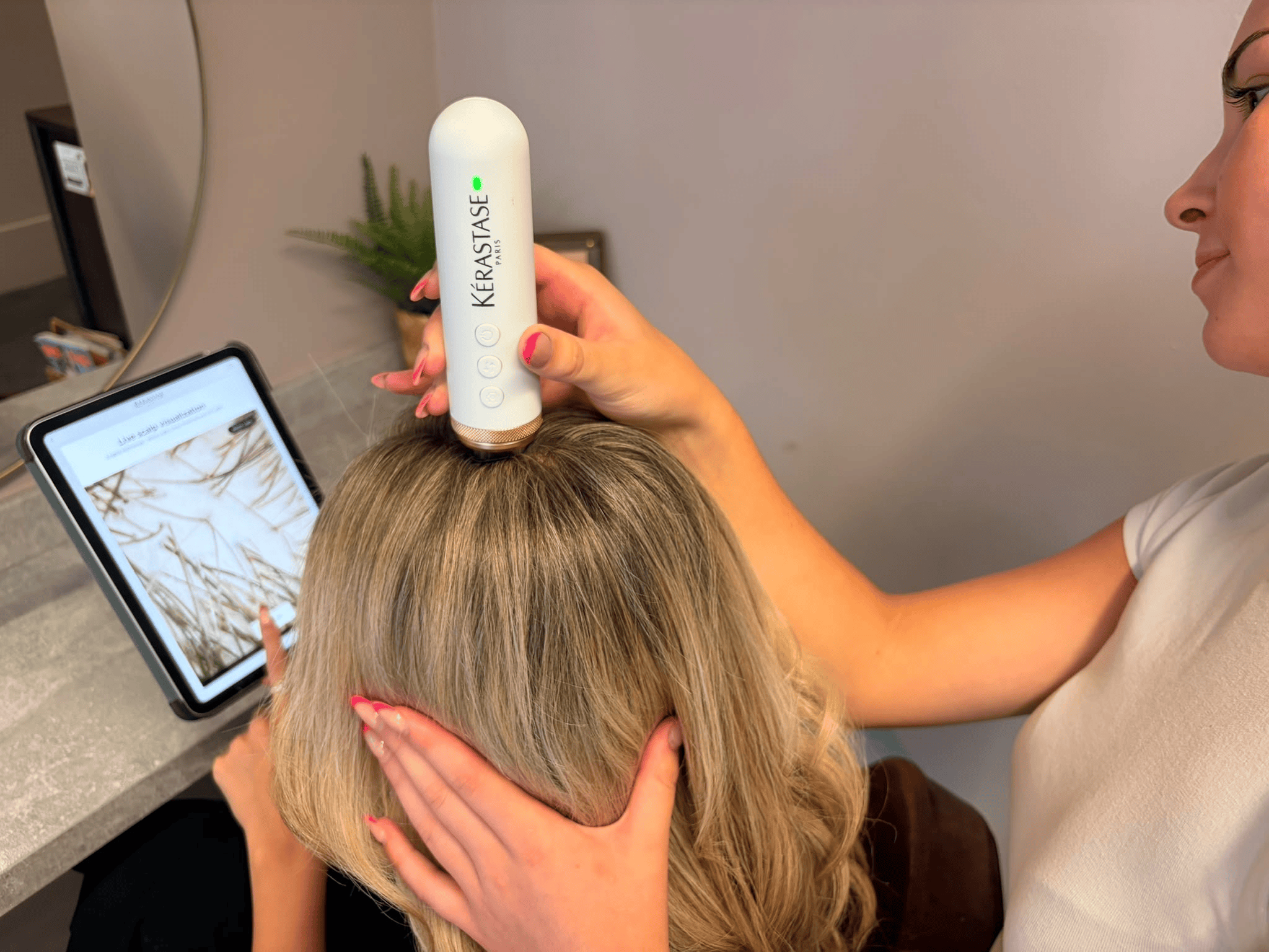 A person with blonde hair is receiving a scalp treatment with a handheld device labeled 'Kérastase Paris' while another person holds their head. A tablet displays an application or image related to scalp health, and a small plant is visible in the background.