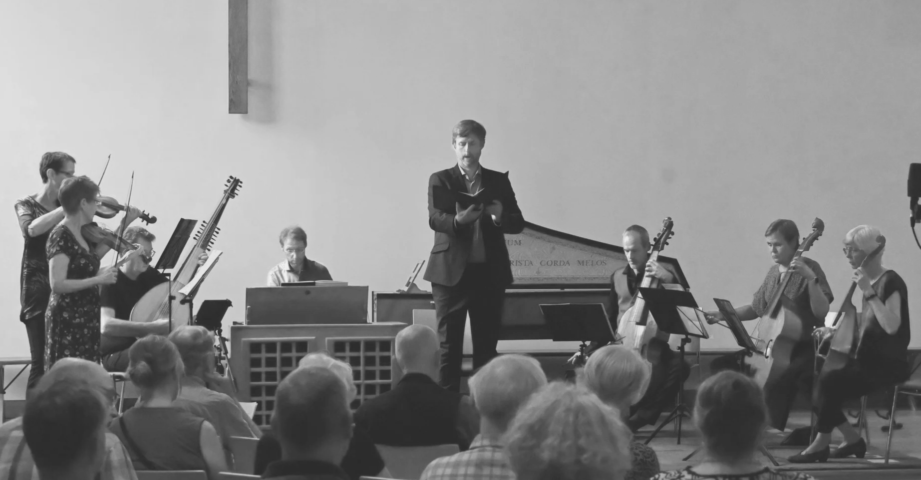 Muzikale uitvoering met een dirigent en meerdere muzikanten die viool, cello, en ander instrument spelen, in een zaal met publiek.