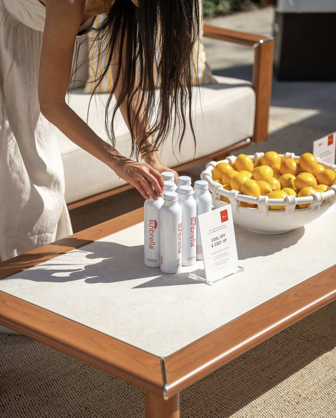 A woman with long hair in a white dress is reaching for bottles on a table outdoors. The table has a large white bowl filled with yellow lemons and a small sign that reads "Cool Off & Cozy Up." There is also a sign with the sun and cloud logo from Su