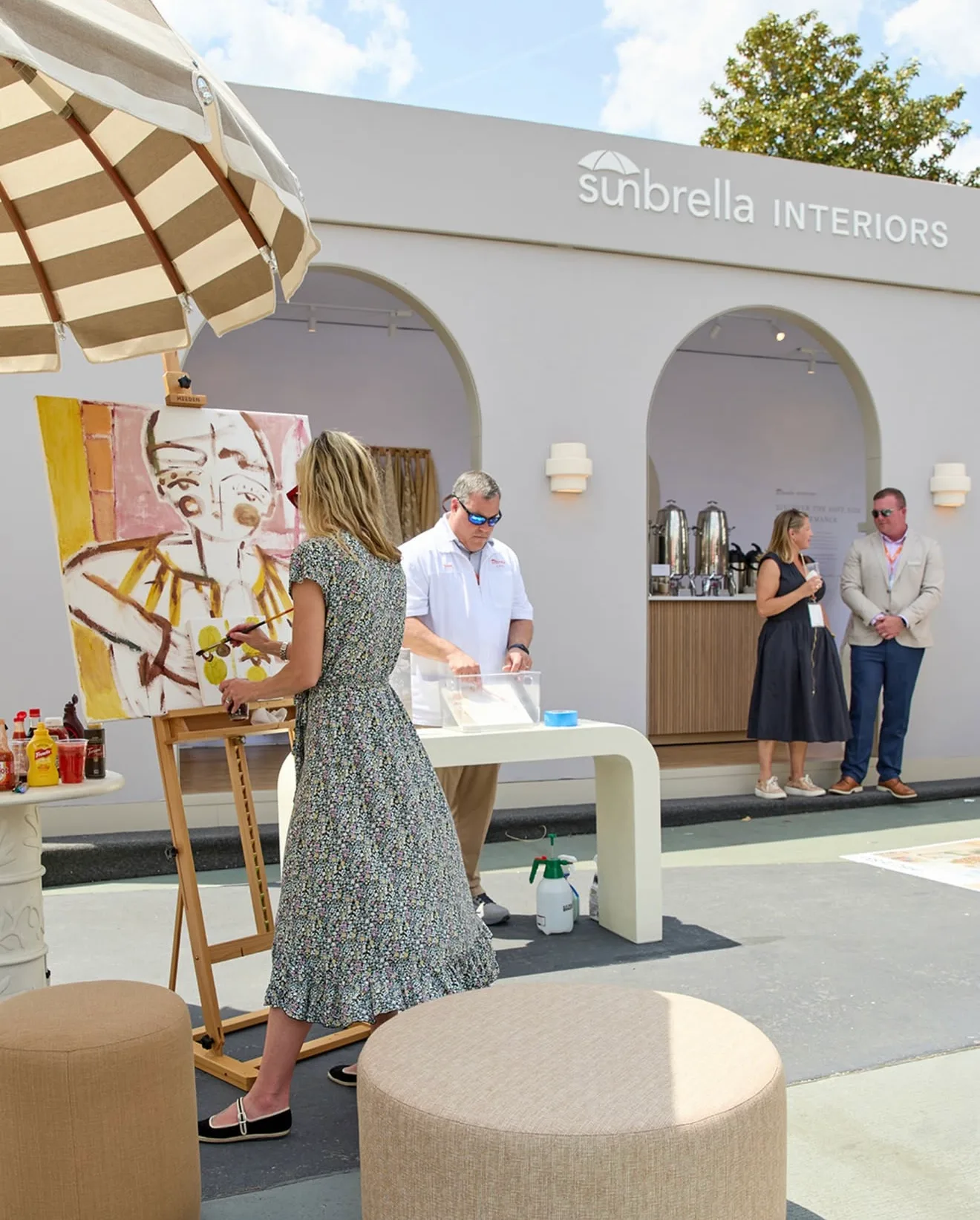 People attending an art event or exhibition outside a store called Sunbella Interiors, with one person painting on a canvas and others socializing near a refreshment station.