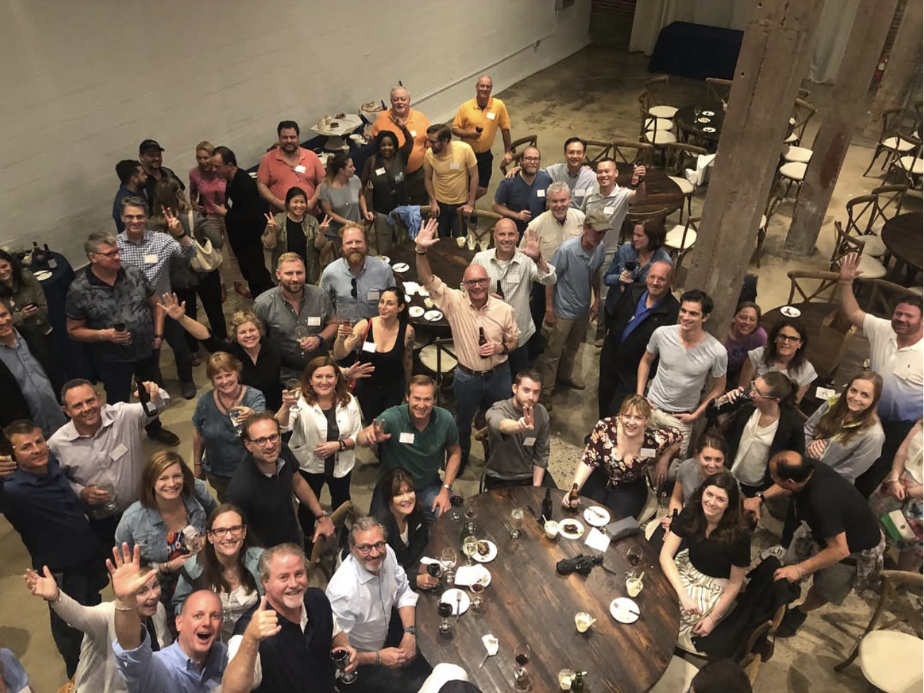 A large group of people at an indoor social gathering or party, many smiling, and some holding drinks, in a space with tables and chairs.
