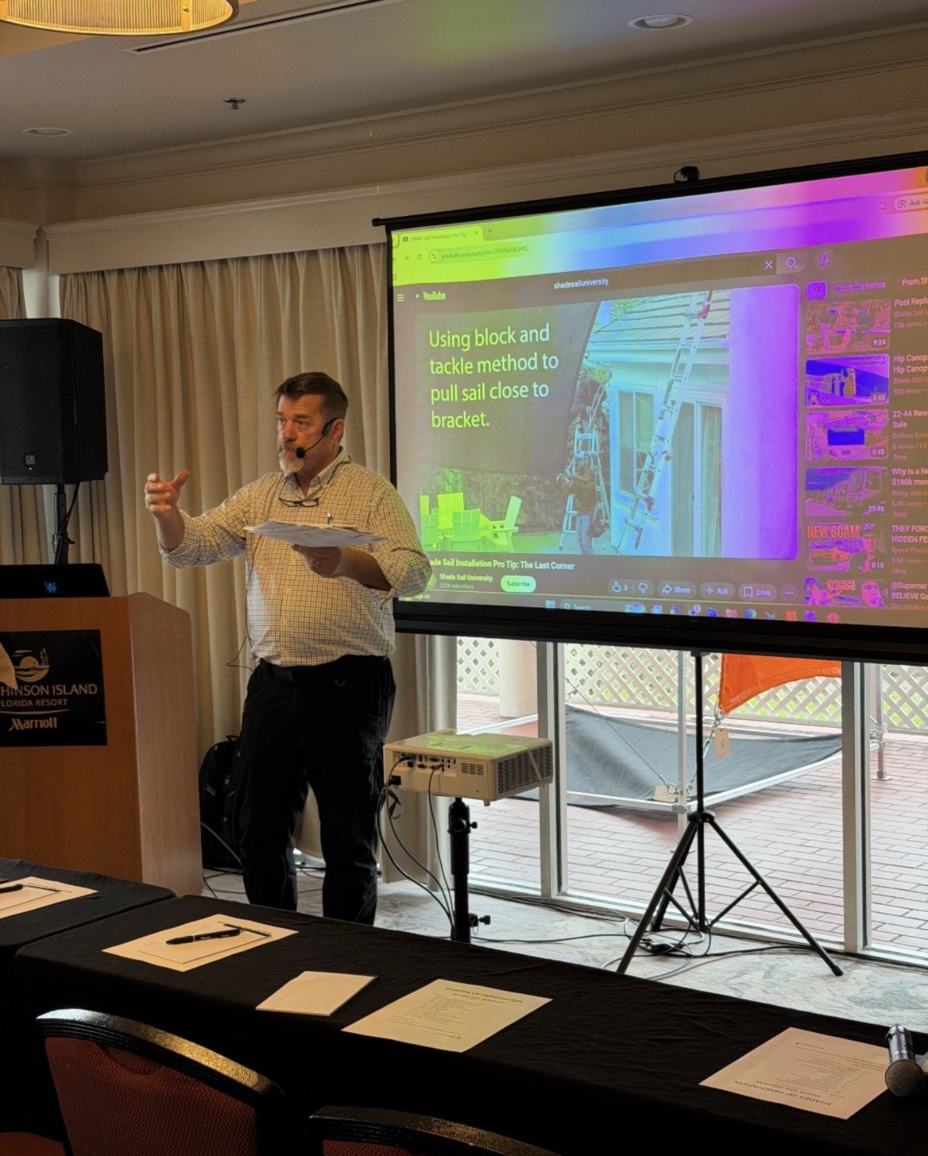 A man giving a presentation in a conference room with a large screen behind him showing a slide on installing a sail using a block and tackle method. The room has beige curtains and a window with a deck outside. There are documents and a microphone o