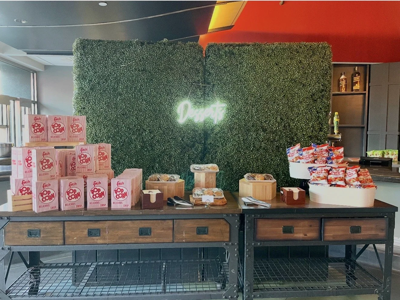 Popcorn boxes stacked on the left side of a wooden table with a green hedge wall and illuminated neon sign that says 'Popcorn' in the background. Snacks and food items arranged on the table.