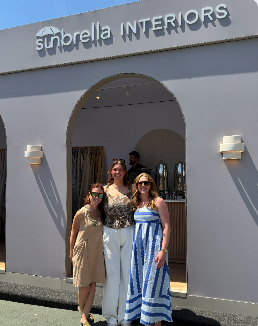 Three women standing outside Sunbrella Interiors store, smiling, with fabric displays and a man in the background.