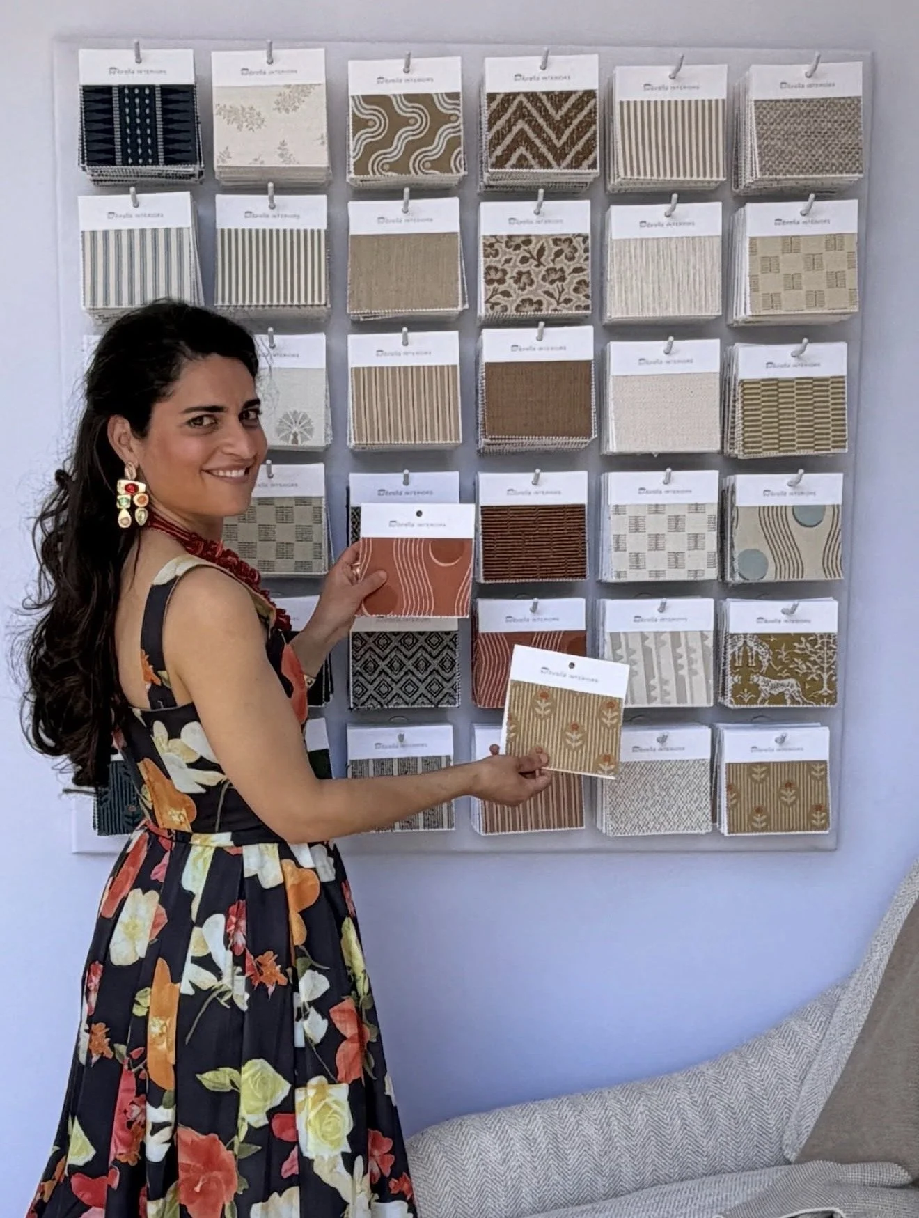 A woman with long dark hair, wearing a floral dress and colorful earrings, stands in front of a wall display of fabric sample swatches, holding two swatches in her hands.