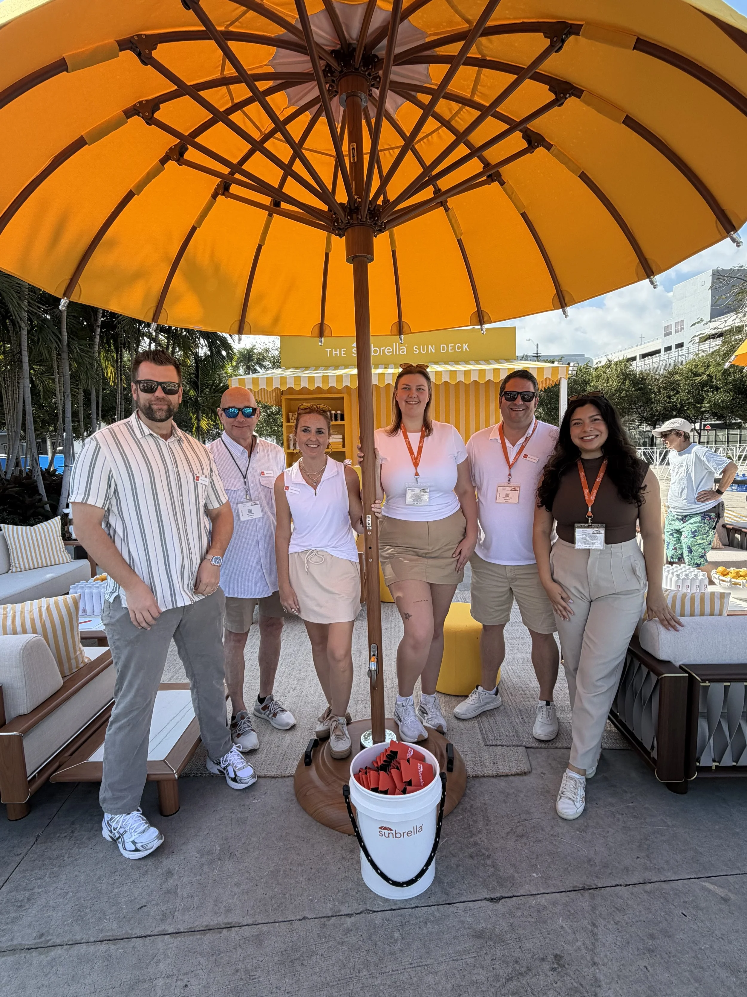 A group of six people standing together under a large yellow umbrella at an outdoor event, with a sunny sky and event signage that says 'The Sunbrella Sun Deck' in the background.