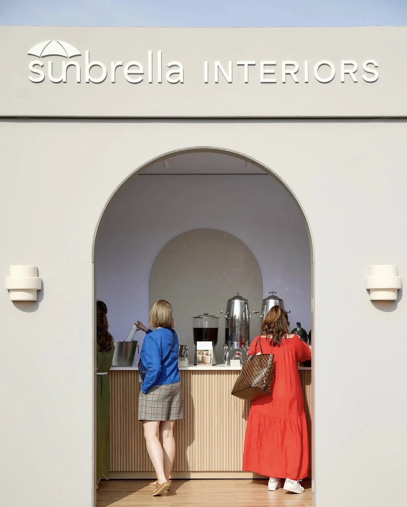 People at a sunbella interiors booth with coffee dispensers and cups, viewed through an arched entrance.
