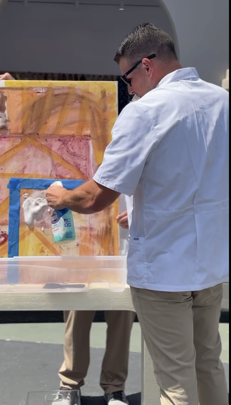 A man with sunglasses and a white shirt cleaning or wiping an acrylic panel with a cloth or sponge. There is an abstract painting in the background.