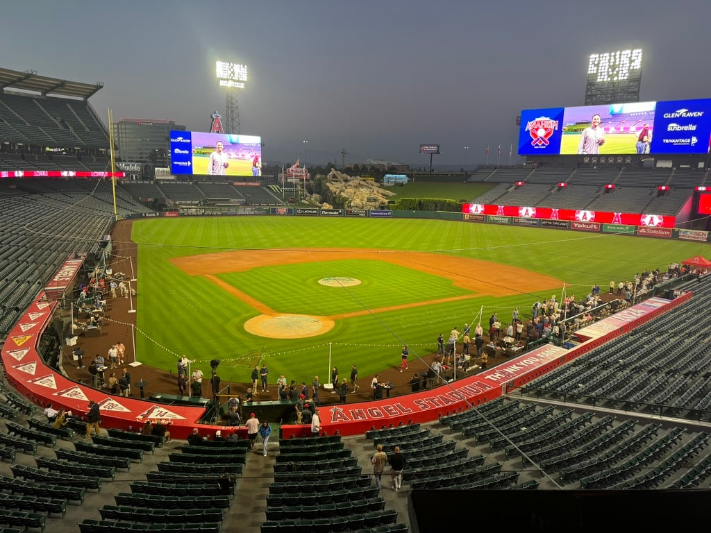 Empty baseball stadium with players practicing on the field, large digital screens displaying advertisements, and a few spectators in the stands.