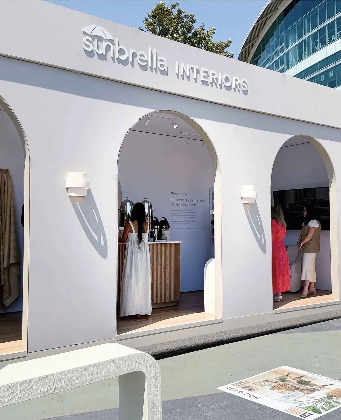 A white outdoor booth with arched openings displaying the Sunbrella Interiors logo. Inside, a woman in a white dress is preparing coffee. Two other women are standing outside the booth, one in a pink dress and the other in a beige vest and white skir