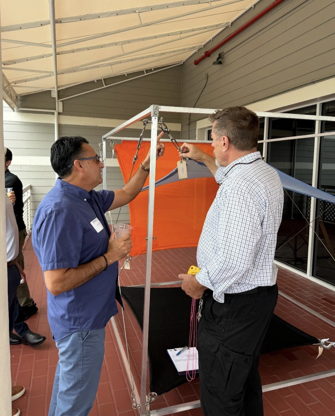 Two men are standing under a shaded patio, working with a small, portable structure supported by a metal frame. One man holds a clear plastic cup, while the other is adjusting a chain or cord attached to the setup. There are other people in the backg