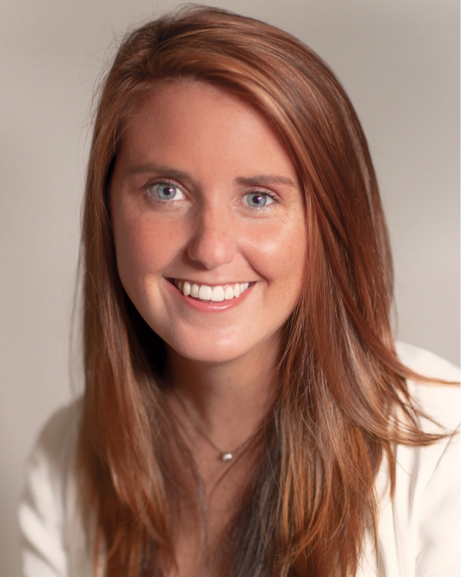 A young woman with long red hair, blue eyes, and a bright smile, wearing a white top and a delicate necklace, against a plain background.