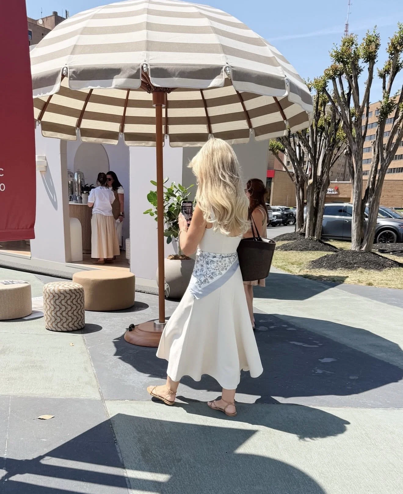 A woman with long blonde hair wearing a white dress takes a photo with her phone under a large striped outdoor umbrella. In the background, two women are inside a small white building, and there are trees, parked cars, and a building with windows beh