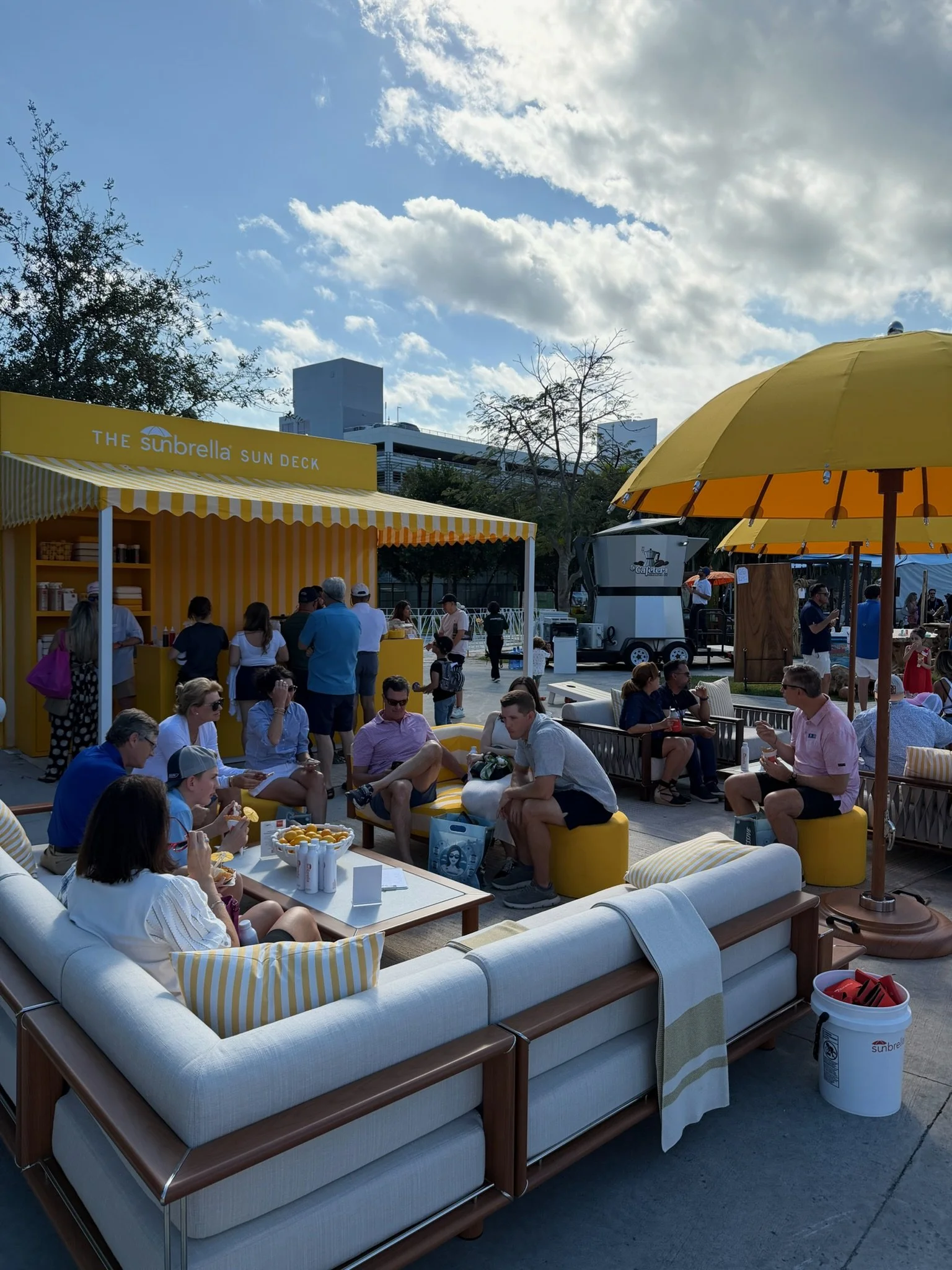 Group of people enjoying outdoor seating at a sunny event with yellow umbrellas and a yellow Sun Deck booth, in an urban setting with trees and modern buildings in the background.