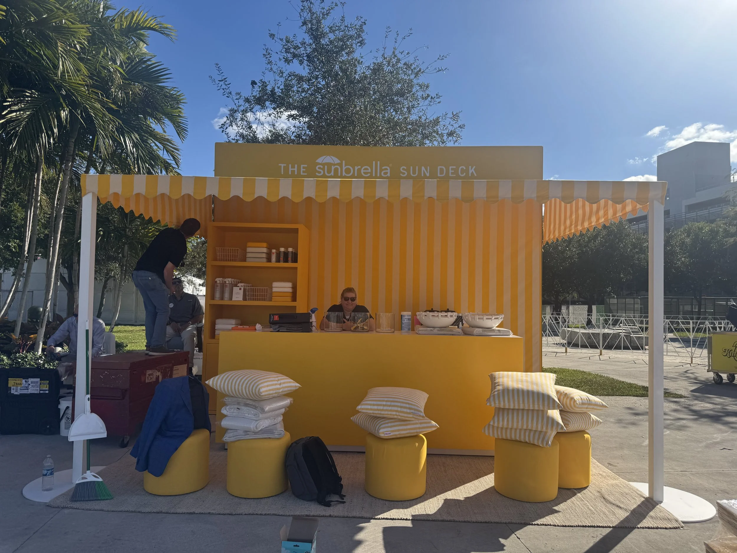 A yellow booth at an outdoor event with a sign that reads 'The Sunbrella Sun Deck'. The booth has striped yellow and white awnings and various pillows and fabrics on display. There are people around, some sitting and some standing, with trees and a b