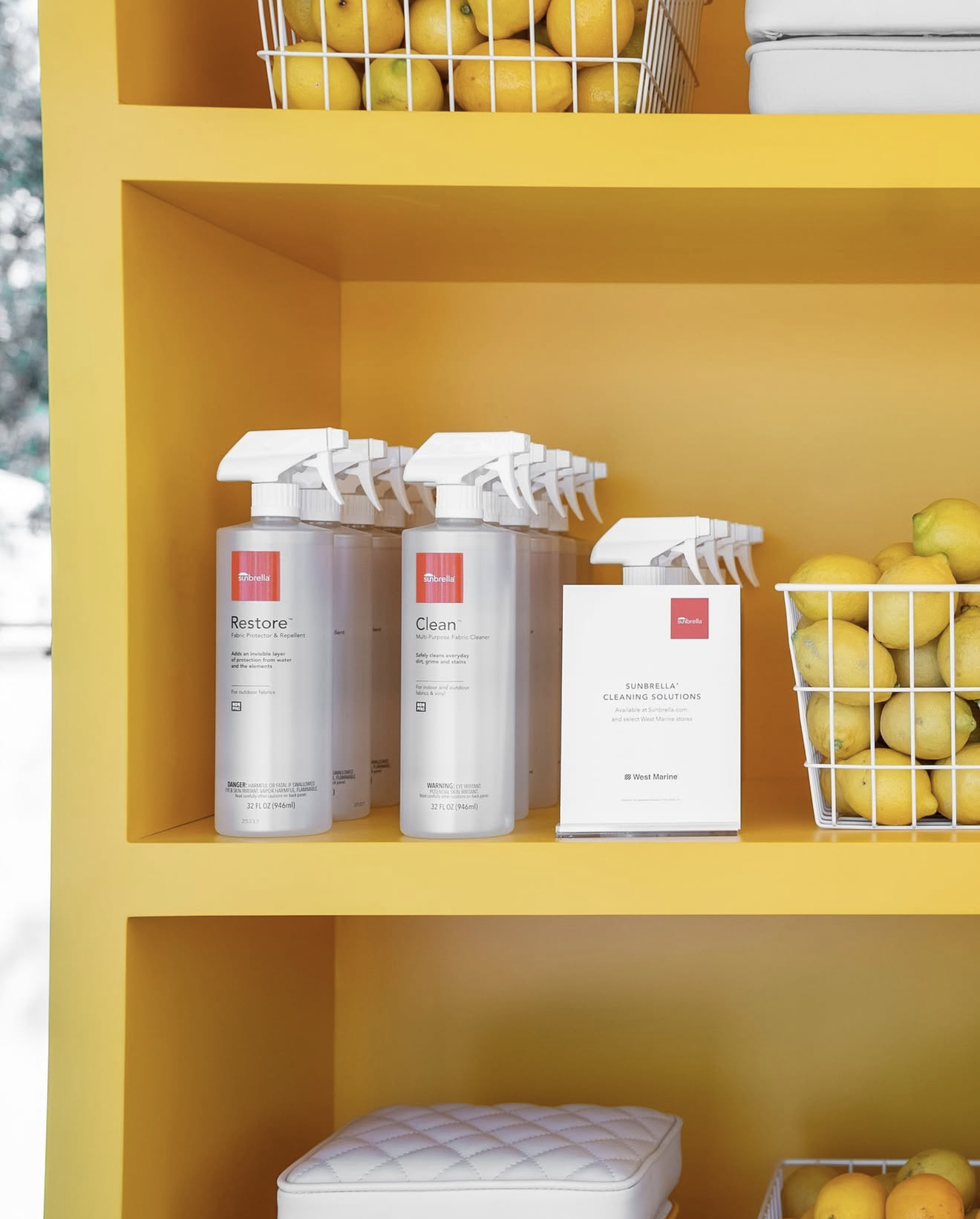 Yellow shelving unit with baskets of lemons, bottles of cleaning solutions labeled 'Restore' and 'Clean,' a white card titled 'Sunbrella Cleaning Solutions,' and a quilted white stool.