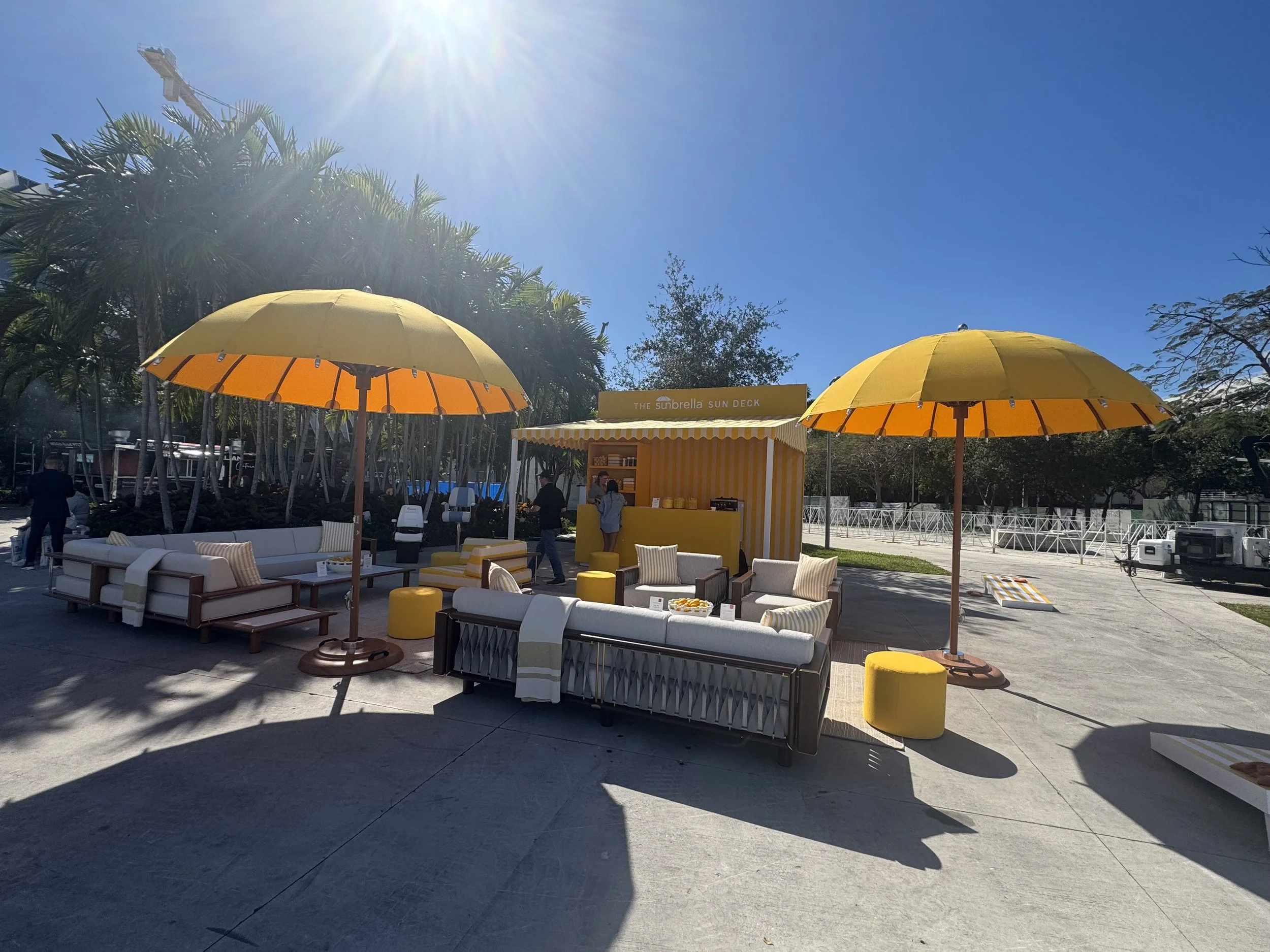 Outdoor seating area with yellow umbrellas, white and yellow furniture, a small food stand labeled 'The Sunbrella Sun Deck', and people walking around on a sunny day with clear blue skies and lush green trees in the background.
