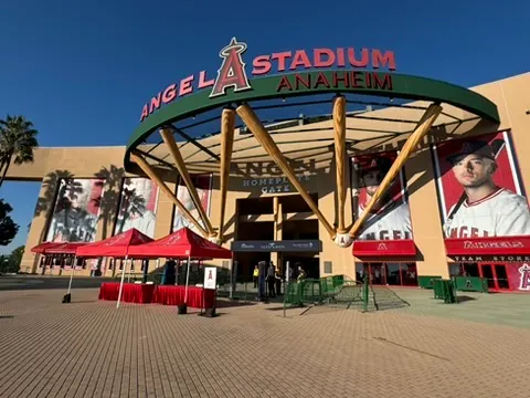 Exterior view of Angel Stadium in Anaheim with the main entrance and large signage, red tents setup outside, clear blue sky, and posters of baseball players.