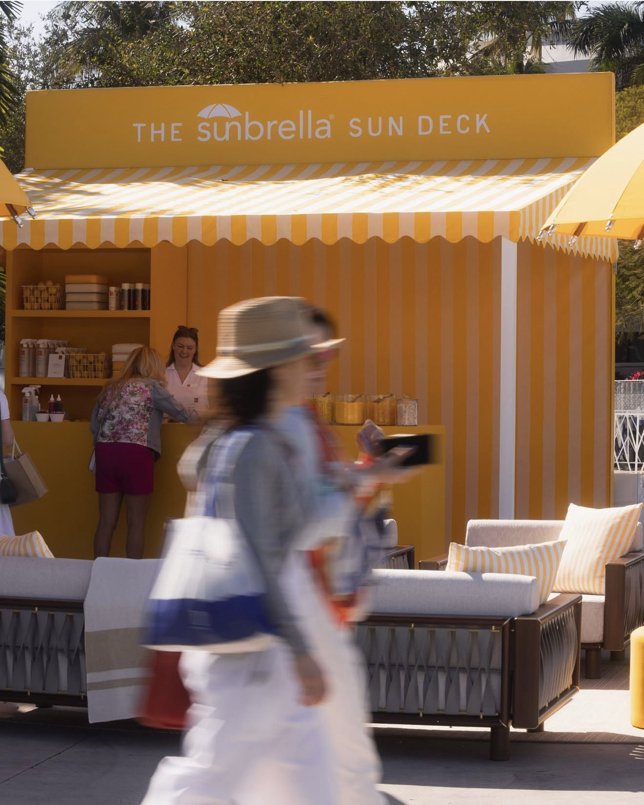 Yellow outdoor café stand with striped yellow and white awning, labeled 'The Sunbrella Sun Deck,' with people walking and a woman ordering at the counter, surrounded by outdoor furniture with striped cushions.