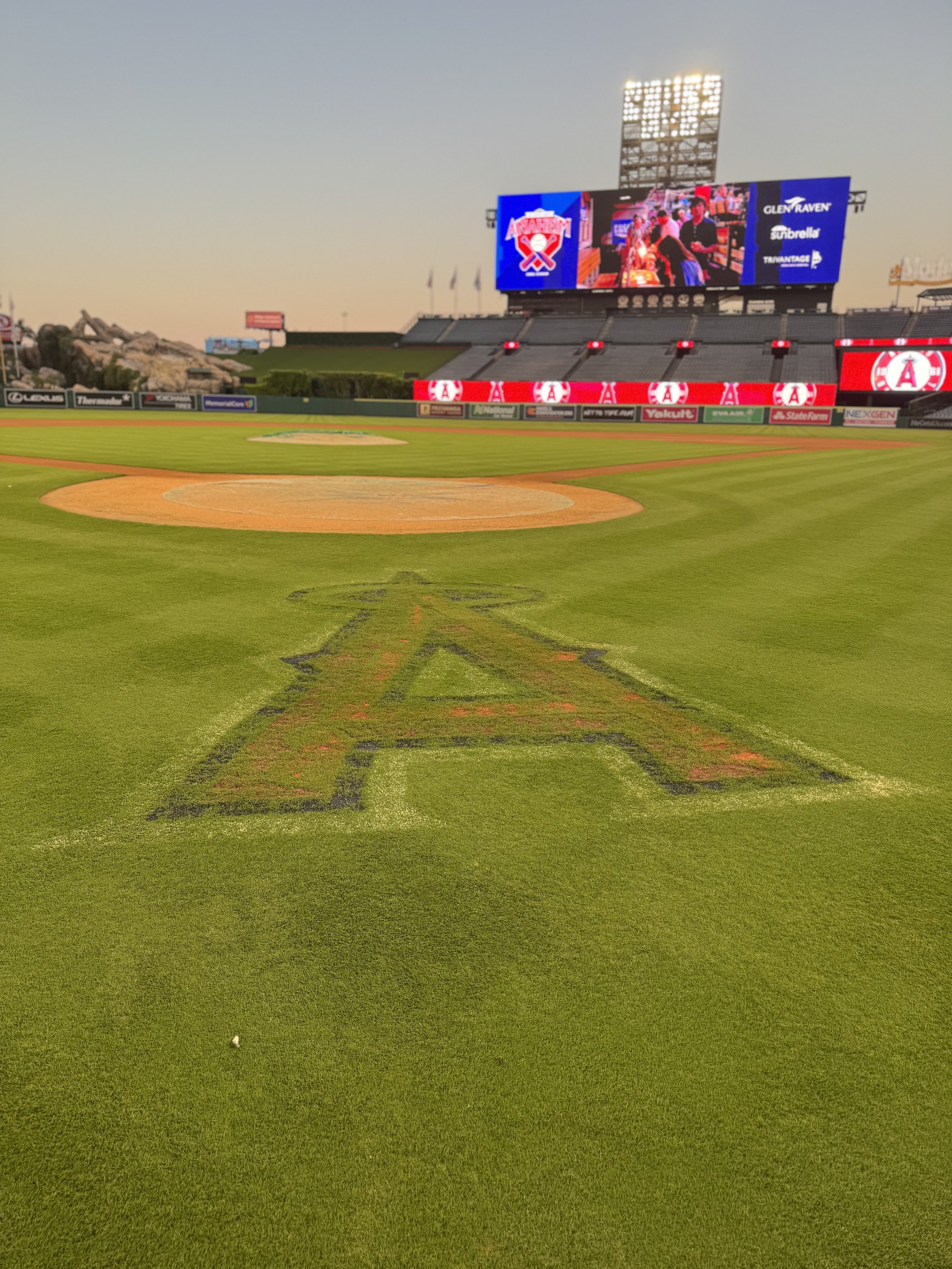 Empty baseball stadium with a field, pitcher's mound, and a large scoreboard displaying ads and the Angels logo at sunset.